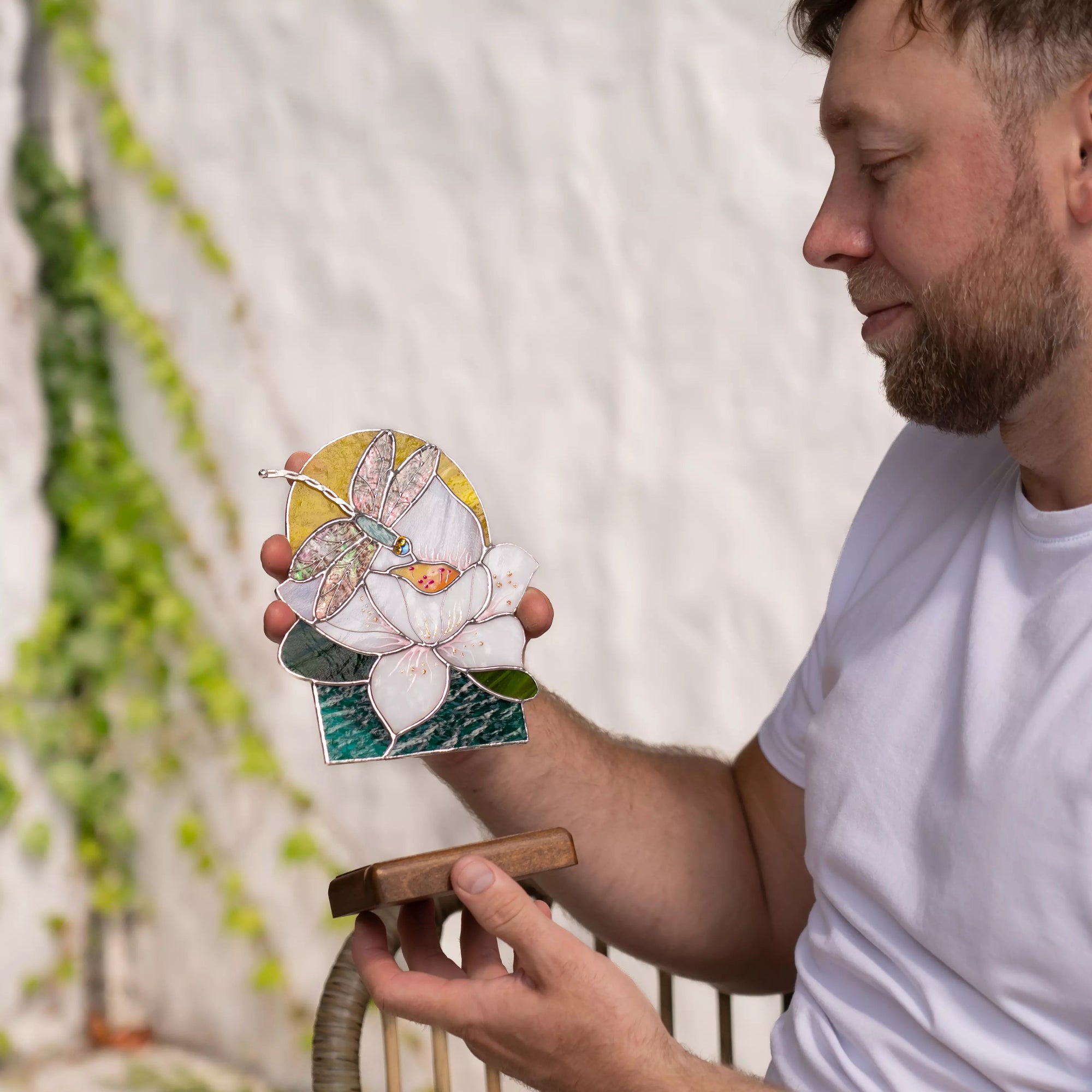 Man holding a small stained glass panel with a dragonfly and floral design outdoors.