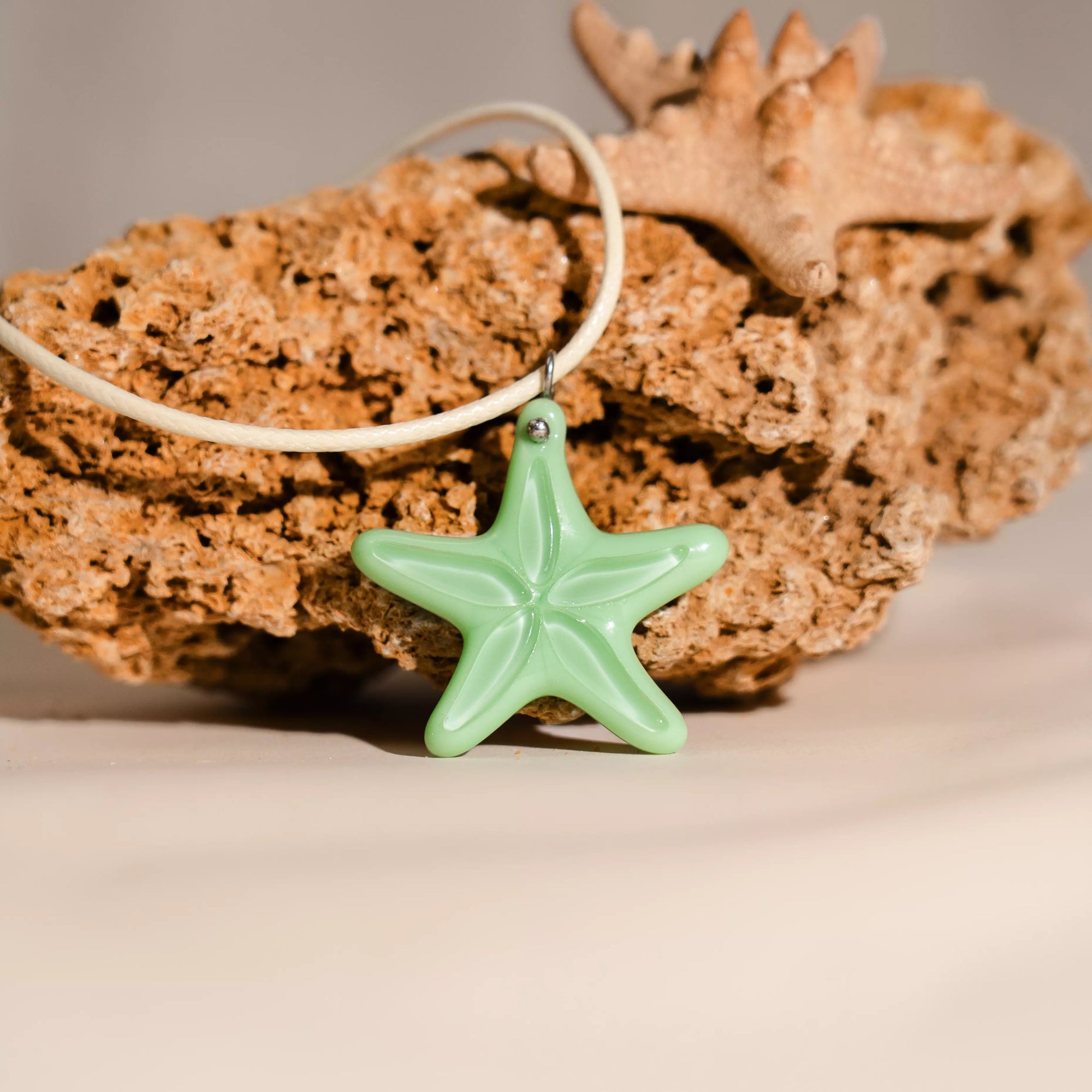 Close-up of mint green starfish pendant on white cord placed against a piece of coral and a small starfish in background.