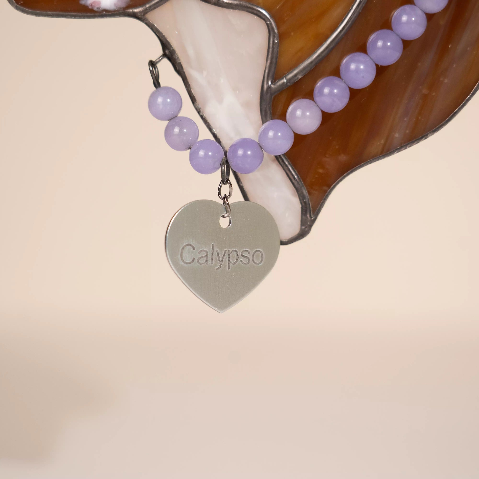 Purple beaded necklace with a silver heart-shaped name tag pendant labeled 'Calypso' on a beige background.