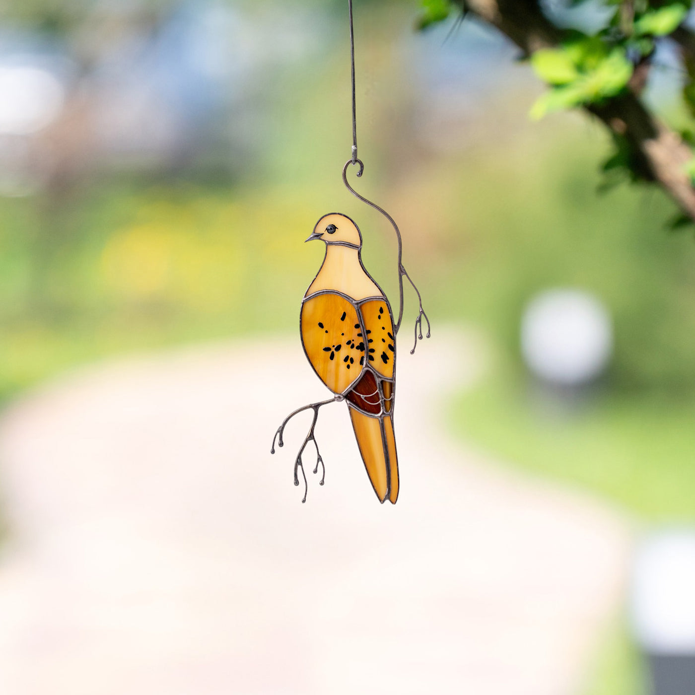 Beige and amber stained glass mourning dove hanging with greenery in background.