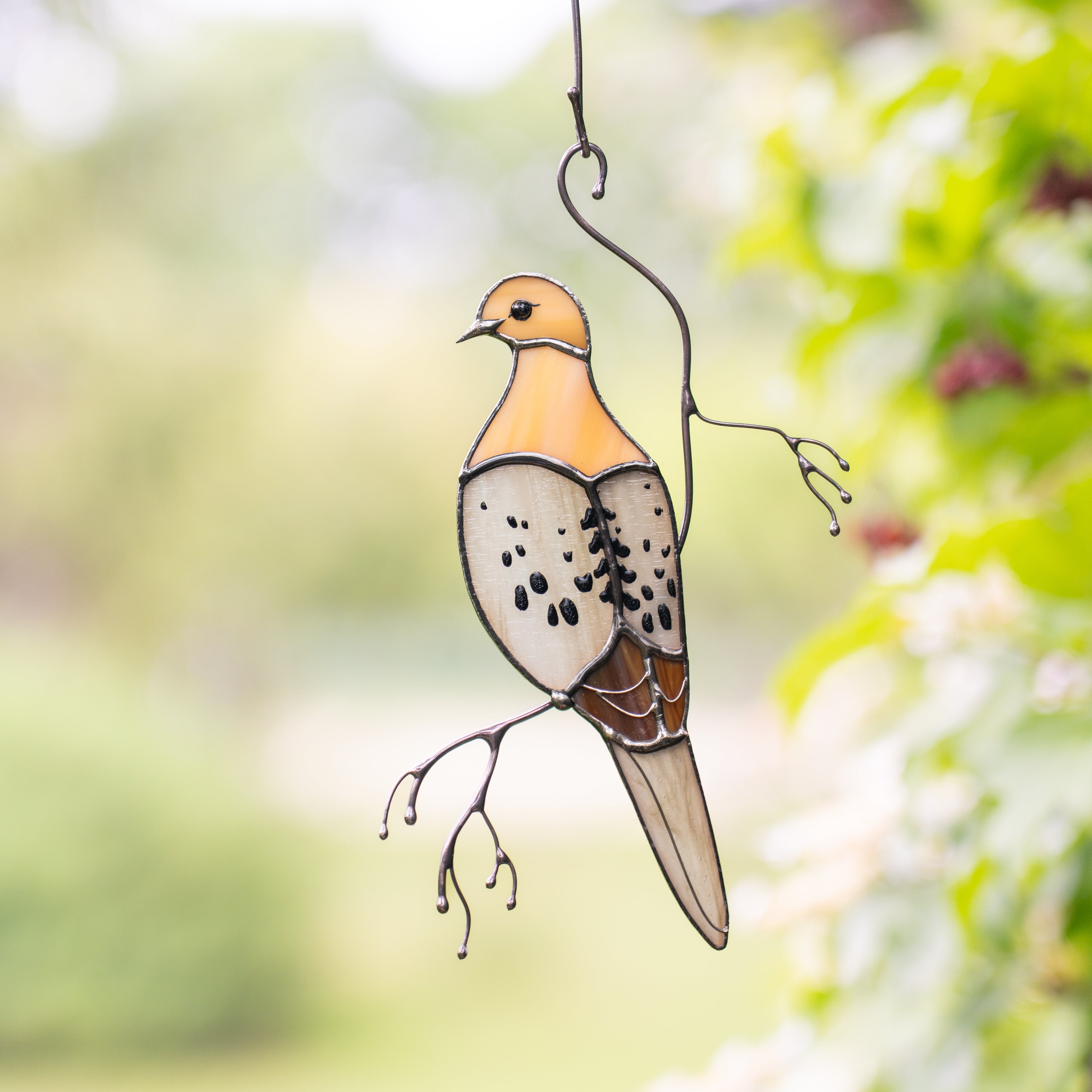Beige and amber stained glass mourning dove hanging with greenery in background.