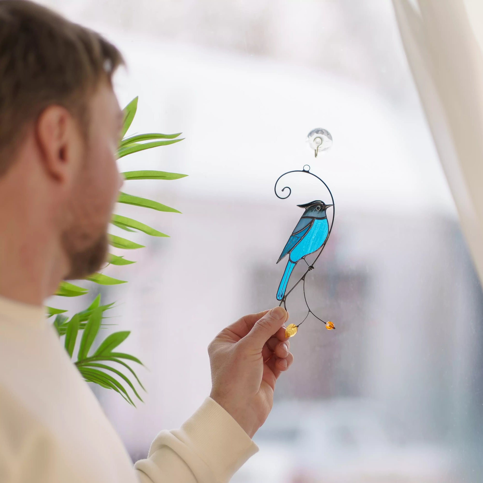 Person holding a blue stellar jay stained glass suncatcher against a blurred indoor background