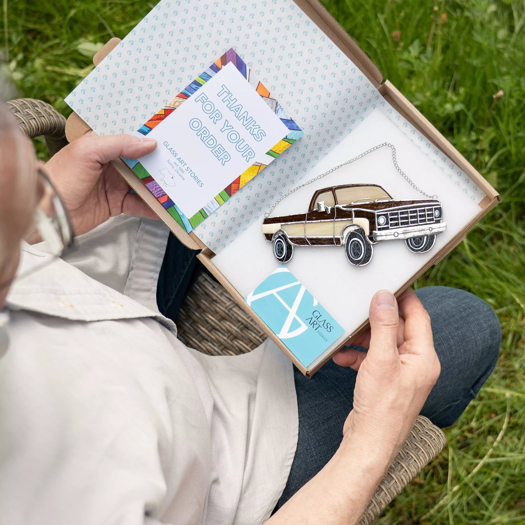 Person holding a box with a truck stained galss suncatcher and a 'Thanks for your order' card on grass.