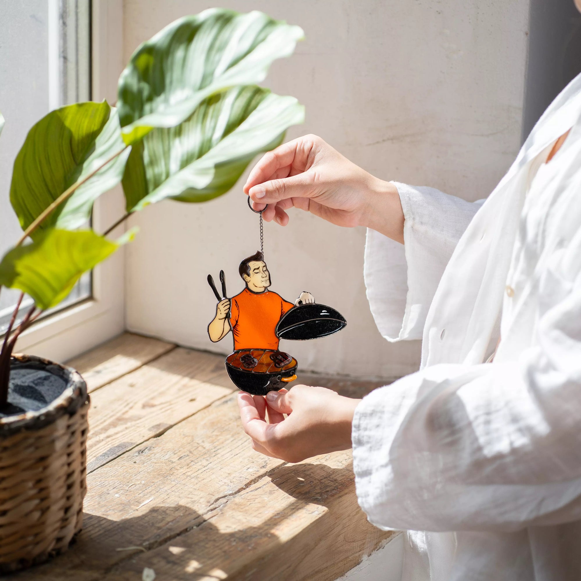 Person holding a stained glasss window hanging barbecue figure by a window with a plant