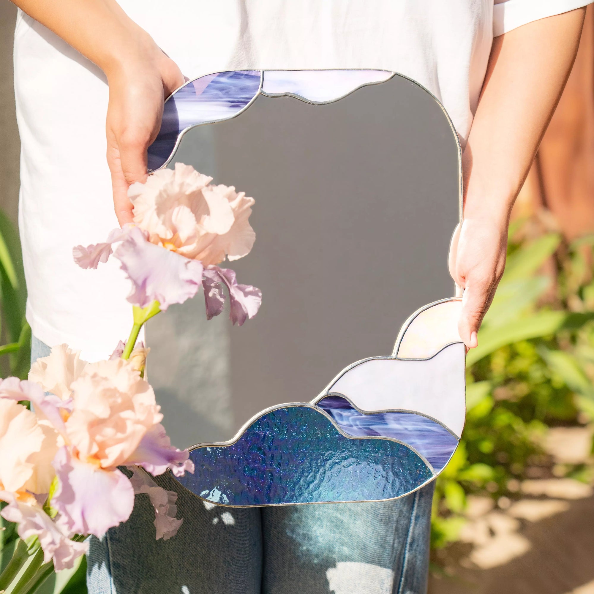 Person holding a stained glass mirror with floral design and flowers in front of it.