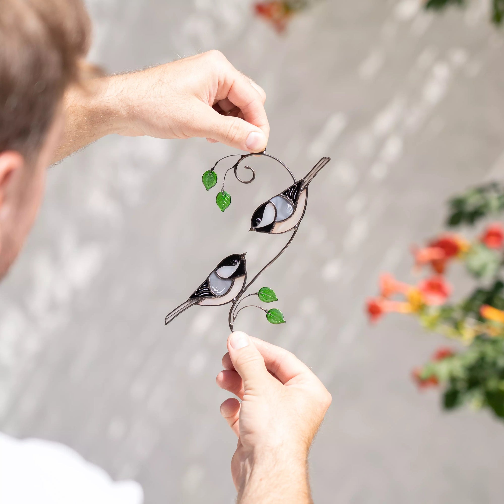 Person holding a decorative stained glass suncatcher with birds and leaves against a  colorful plants with flowers