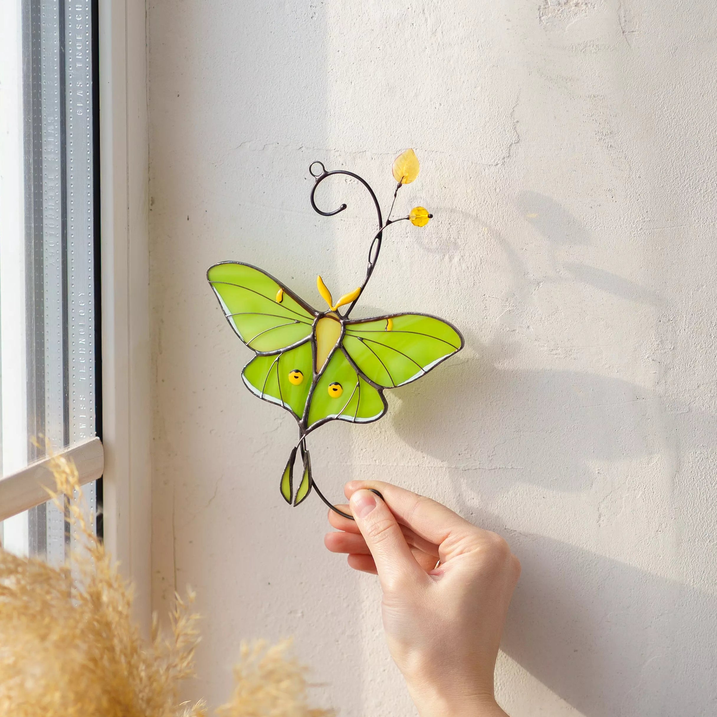 Person holding a green butterfly stained glass window hanging against a light wall.