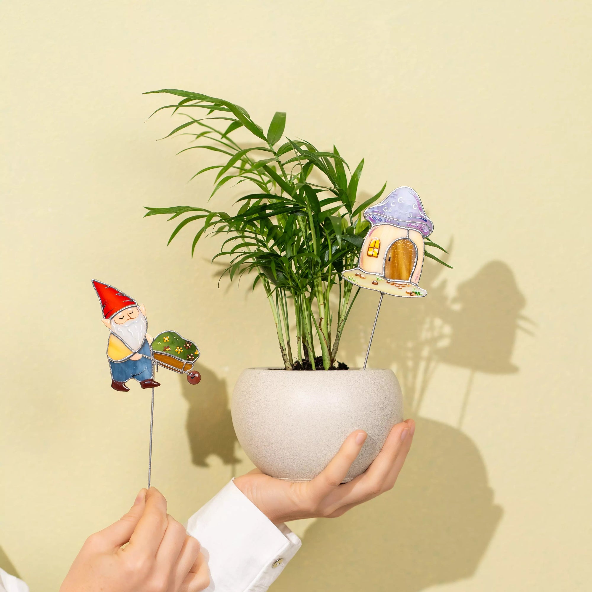 Hands holding a potted plant with a small gnome and fairy house decoration against a yellow background