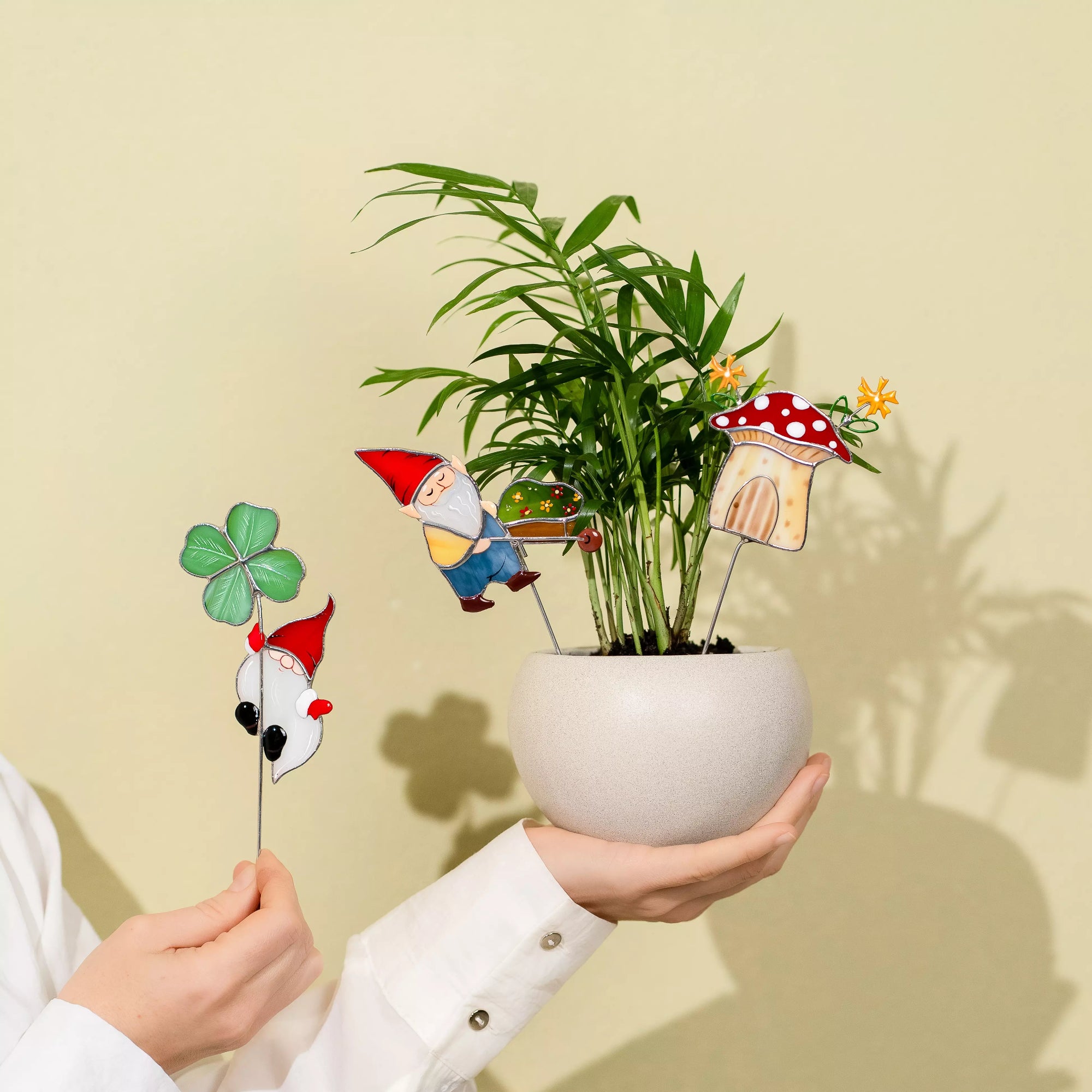Hands holding a potted plant with decorative garden gnome and mushroom stakes against a beige background
