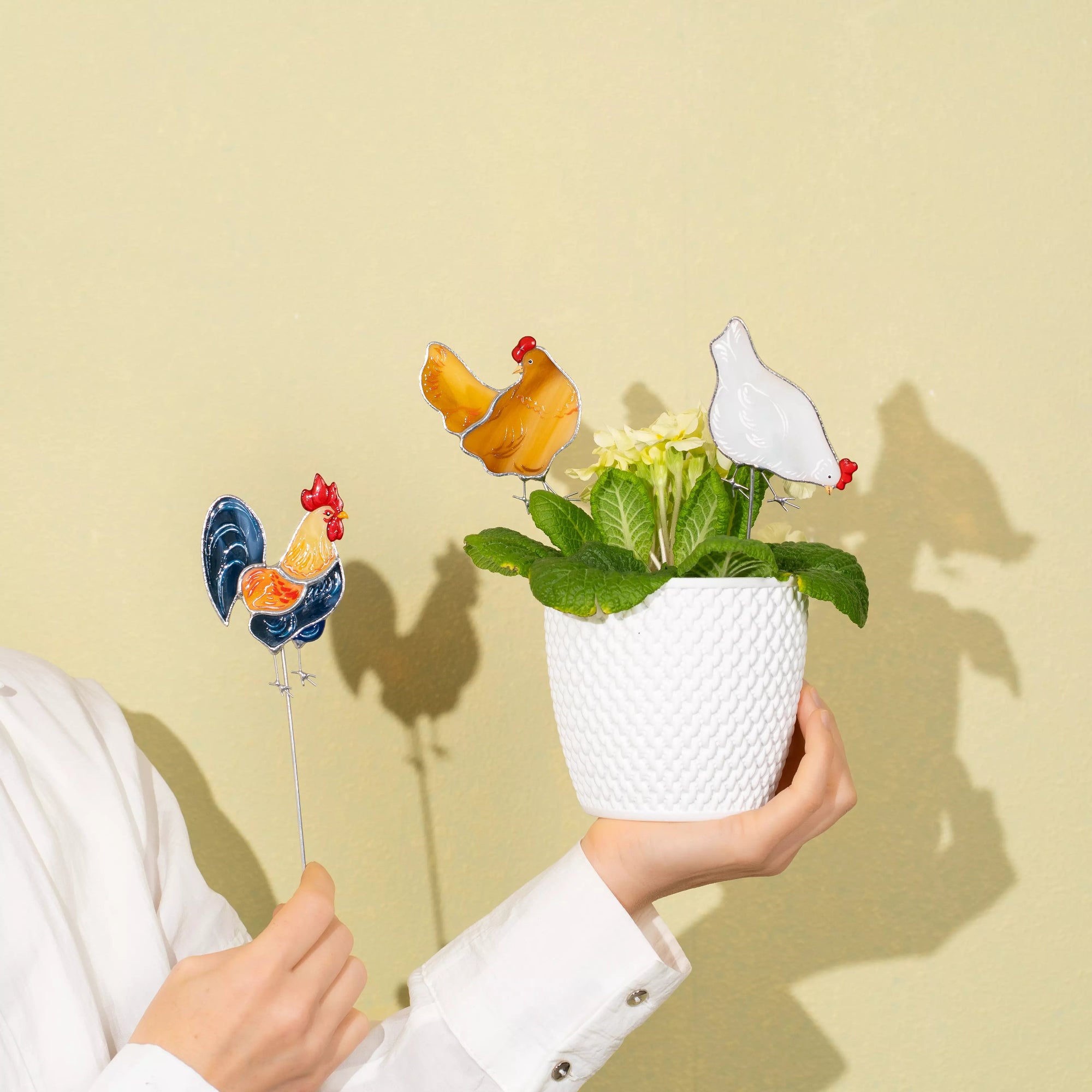Person holding a small potted plant with stained glass chicken plant stakes on skewers against a beige background