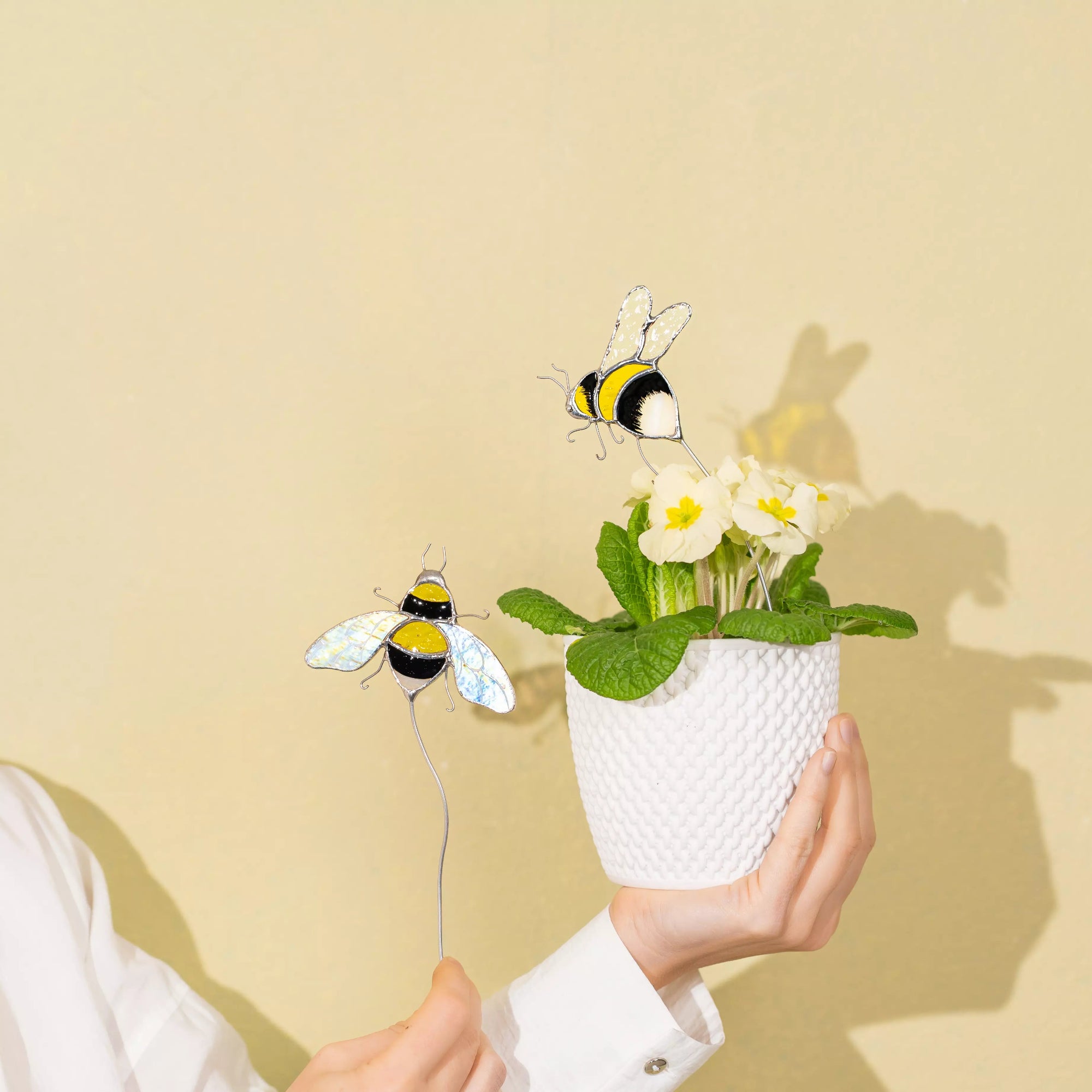 Hands holding a small white pot with flowers and stained glass bee plant stakes against a beige background