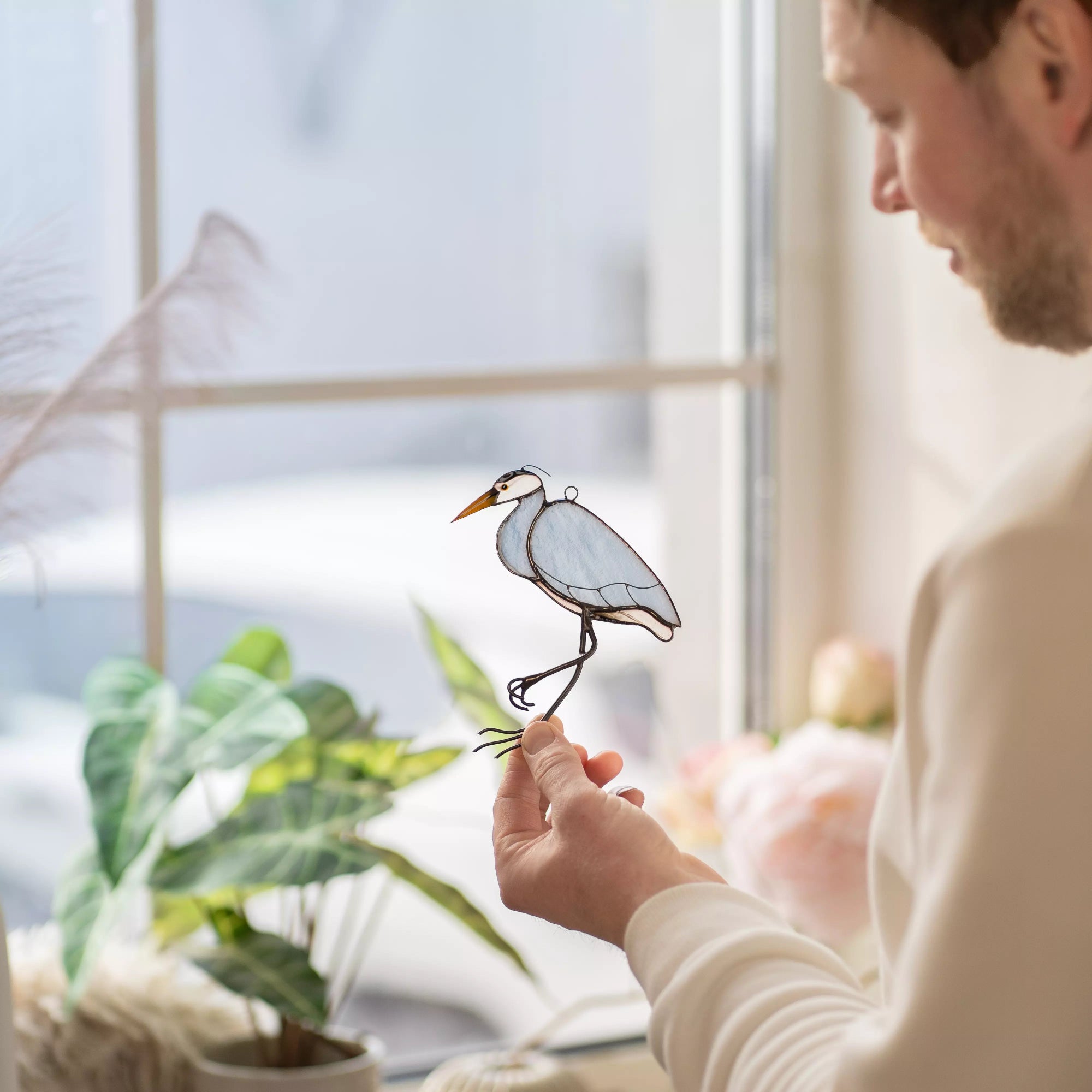 Man holding a stained glass heron bird in front of a window with plants and decor.