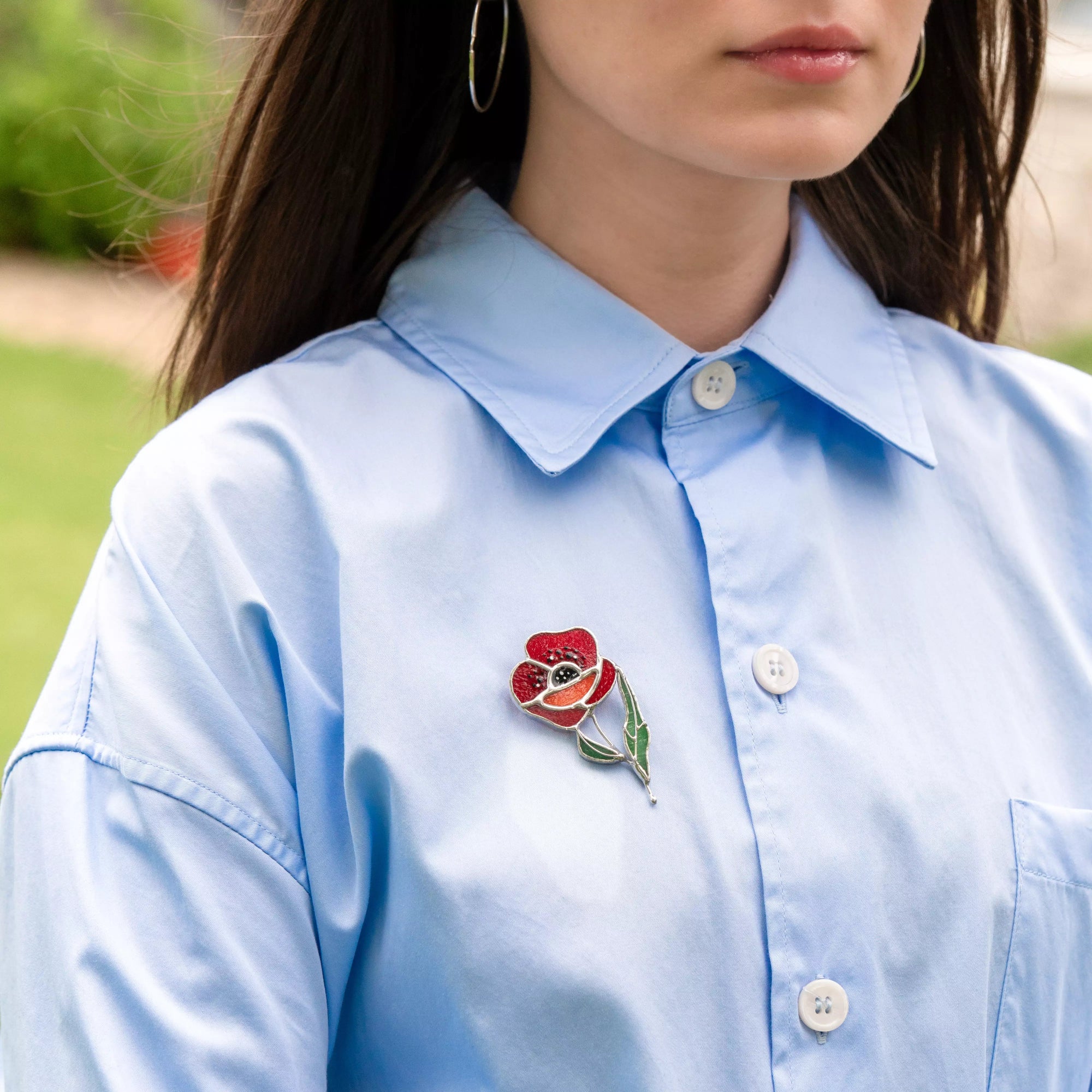 Person wearing a light blue shirt with a red poppy stained glass brooch outdoors