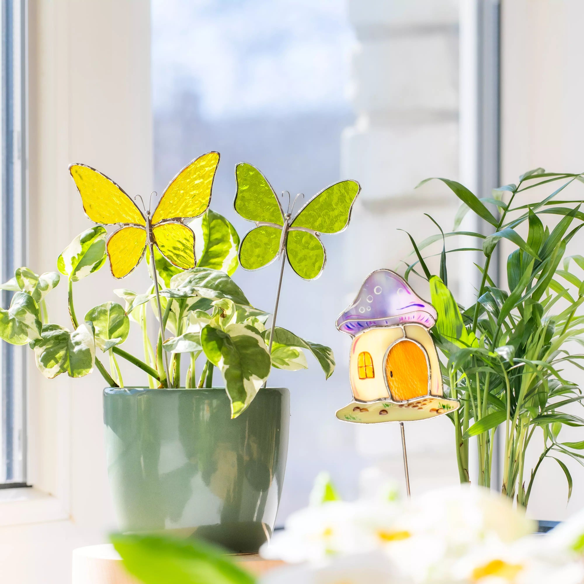A bright lifestyle shot of a yellow butterfly, green butterfly, and mushroom house stake placed in potted plants on a white surface with soft bokeh lighting in the background.