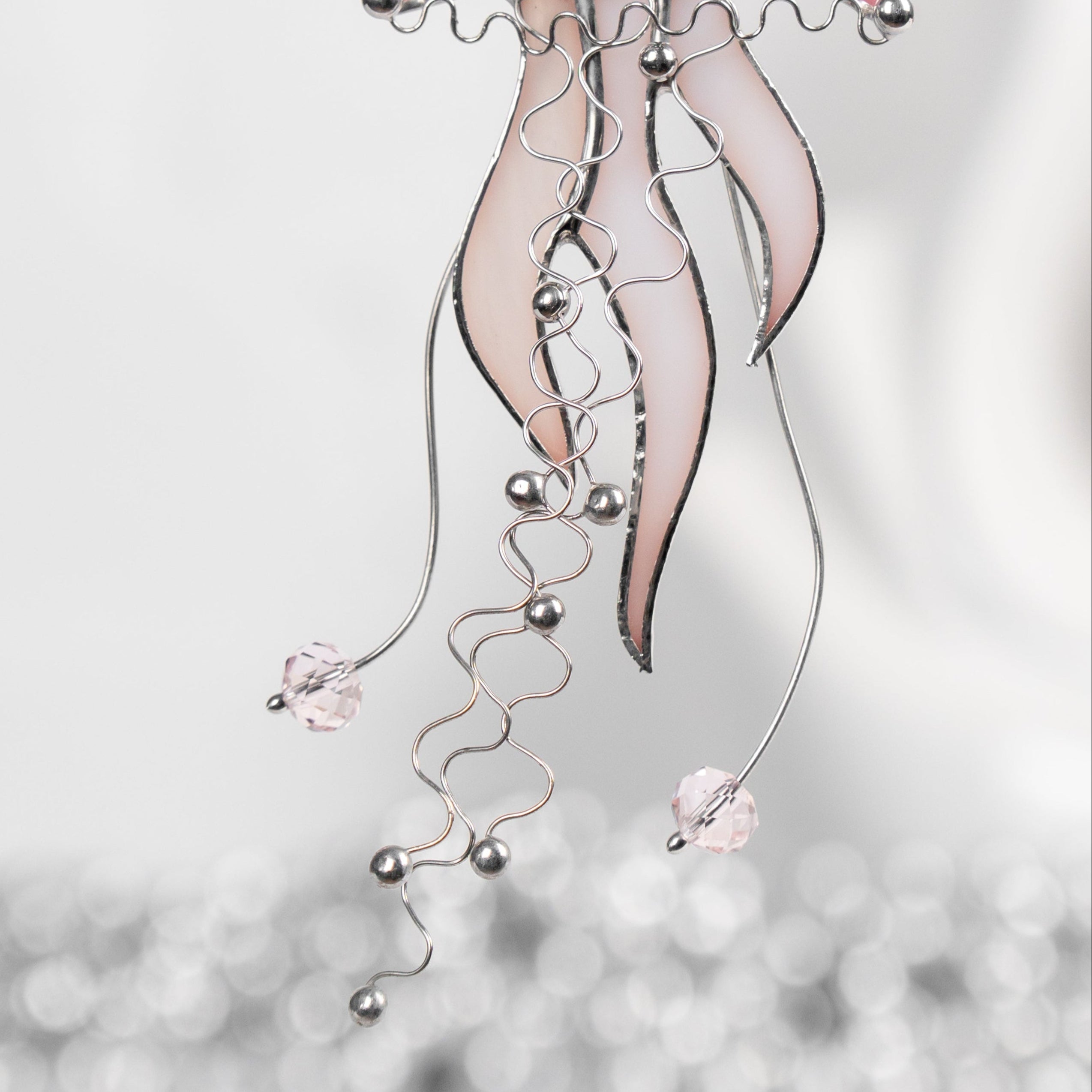 A close-up of delicate pink stained glass jellyfish with swirling wire tentacles and beads, hanging in a bright, softly blurred interior.