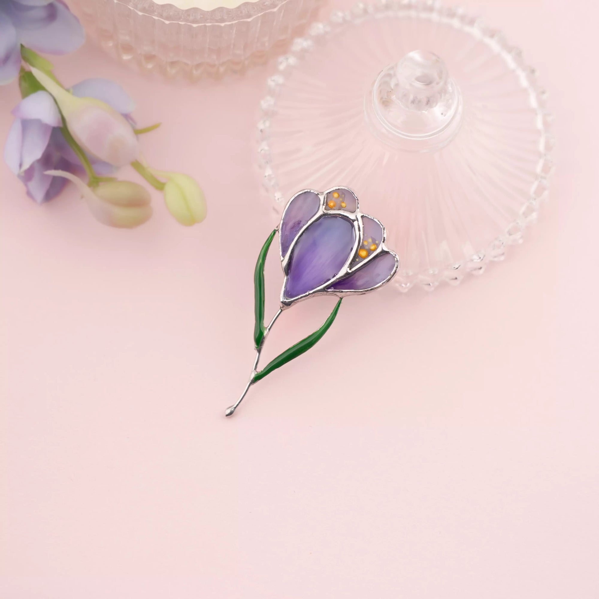 Purple stained glass crocus brooch arranged on a soft pink background with delicate decor, highlighting glass texture and color.