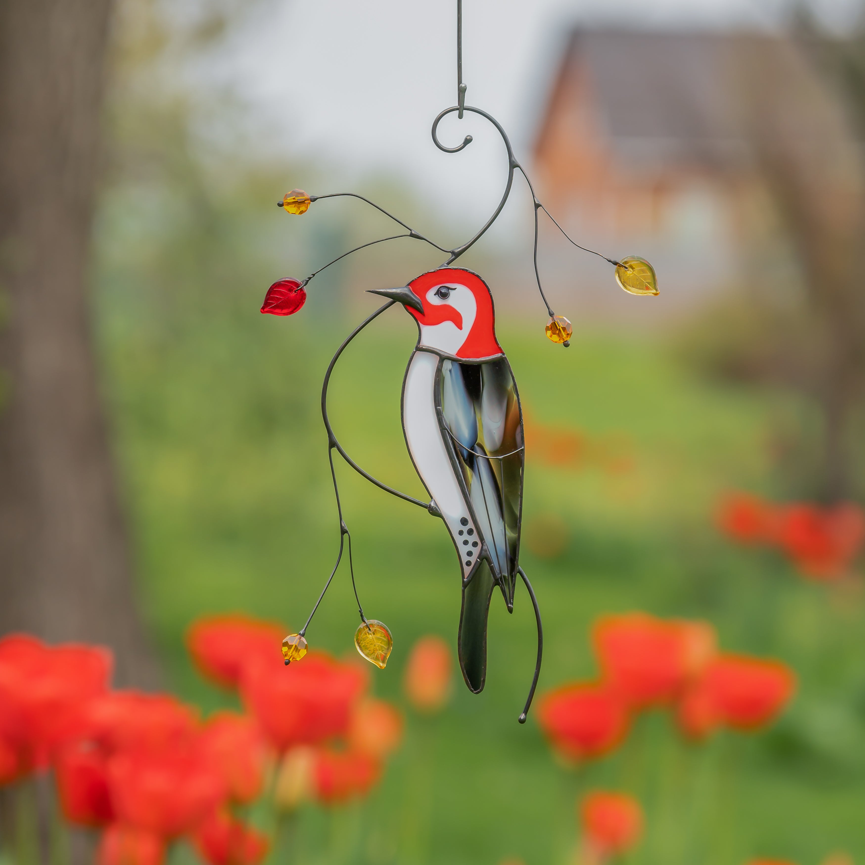 Stained glass woodpecker with a red head and amber leaves hanging outdoors with red tulips in the background.