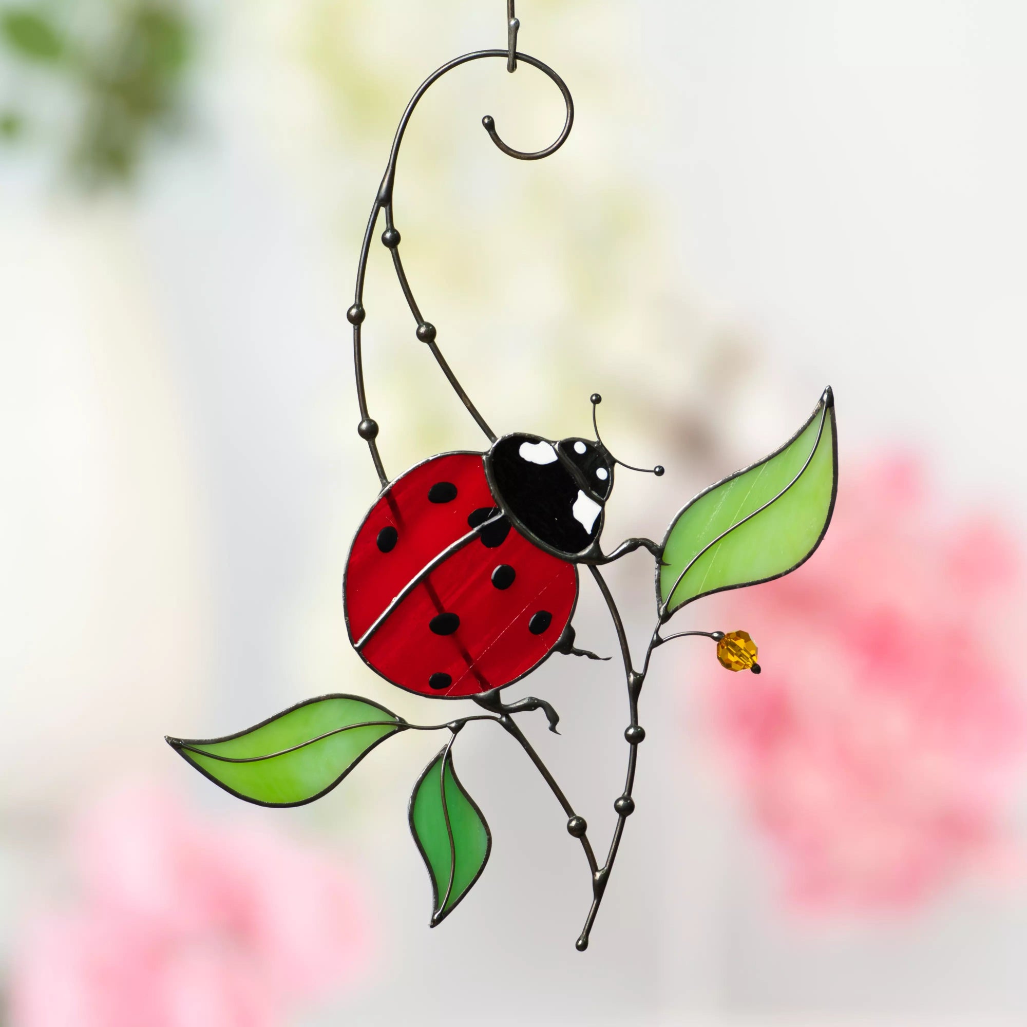Ladybug stained glass hanging with red wings and black spots, resting on a metal branch with green leaves and a yellow crystal accent.
