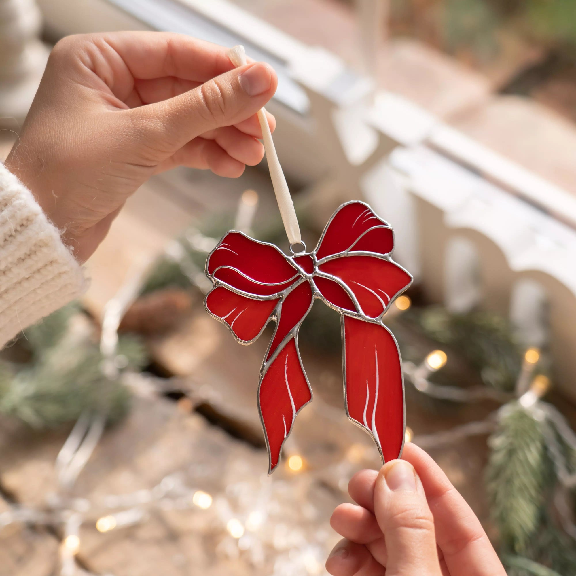 Red bow stained glass ornament held by a person with blurred Christmas lights and decorations in the background