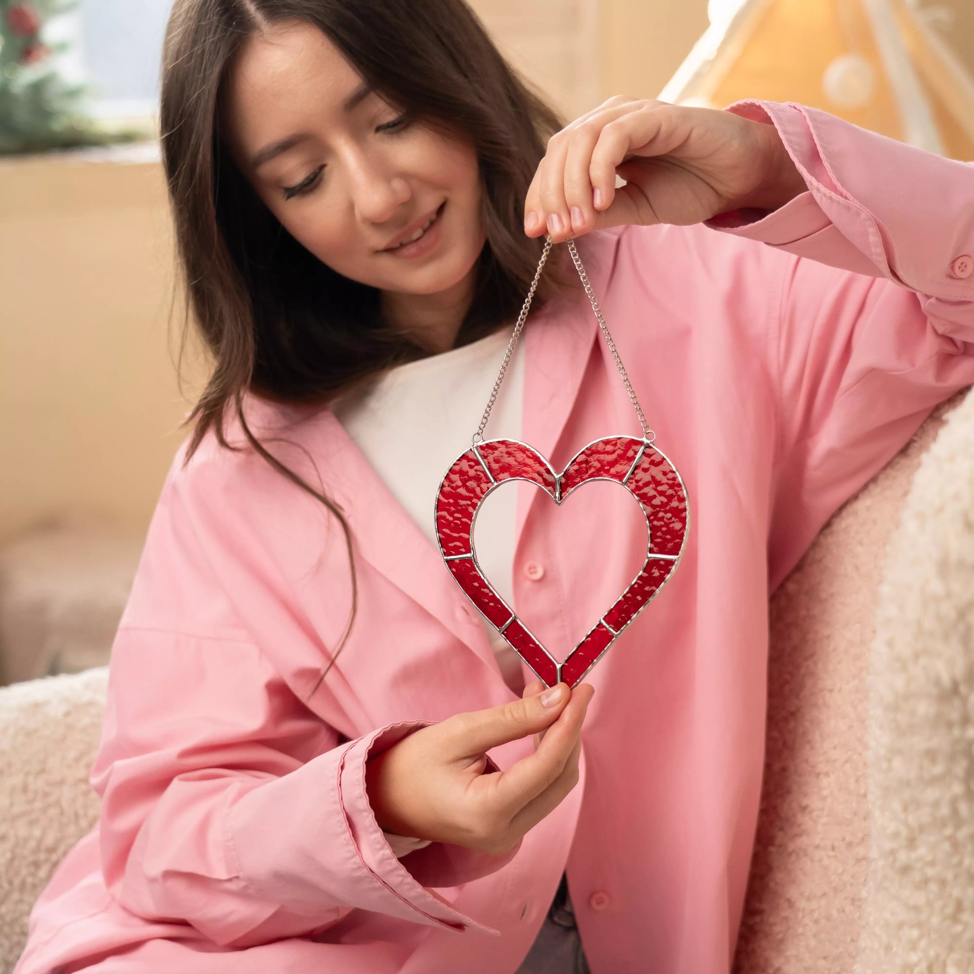 Woman holding a red stained glass heart suncatcher in a cozy interior.