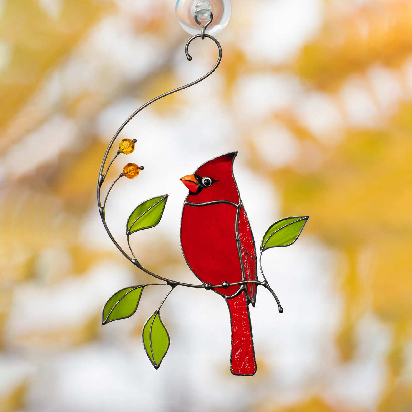 Bright red stained glass cardinal bird perched on a metal branch with green leaves and orange beads, hanging on window