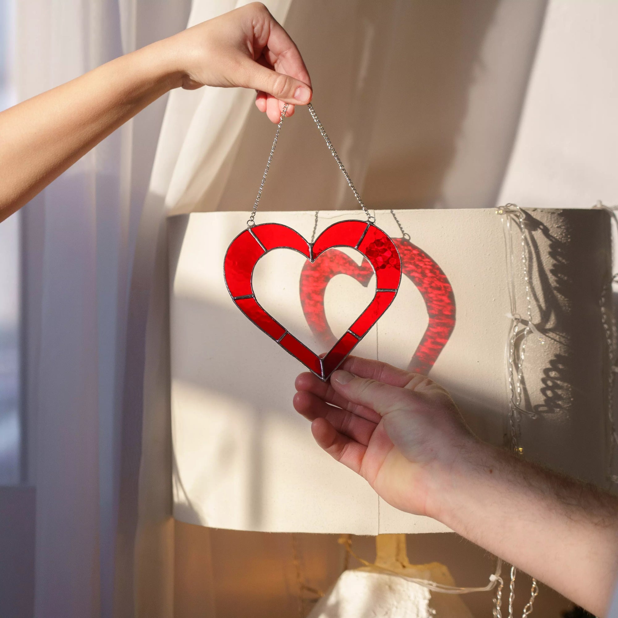 Red stained glass heart suncatcher held by two hands, symbolizing love and connection.