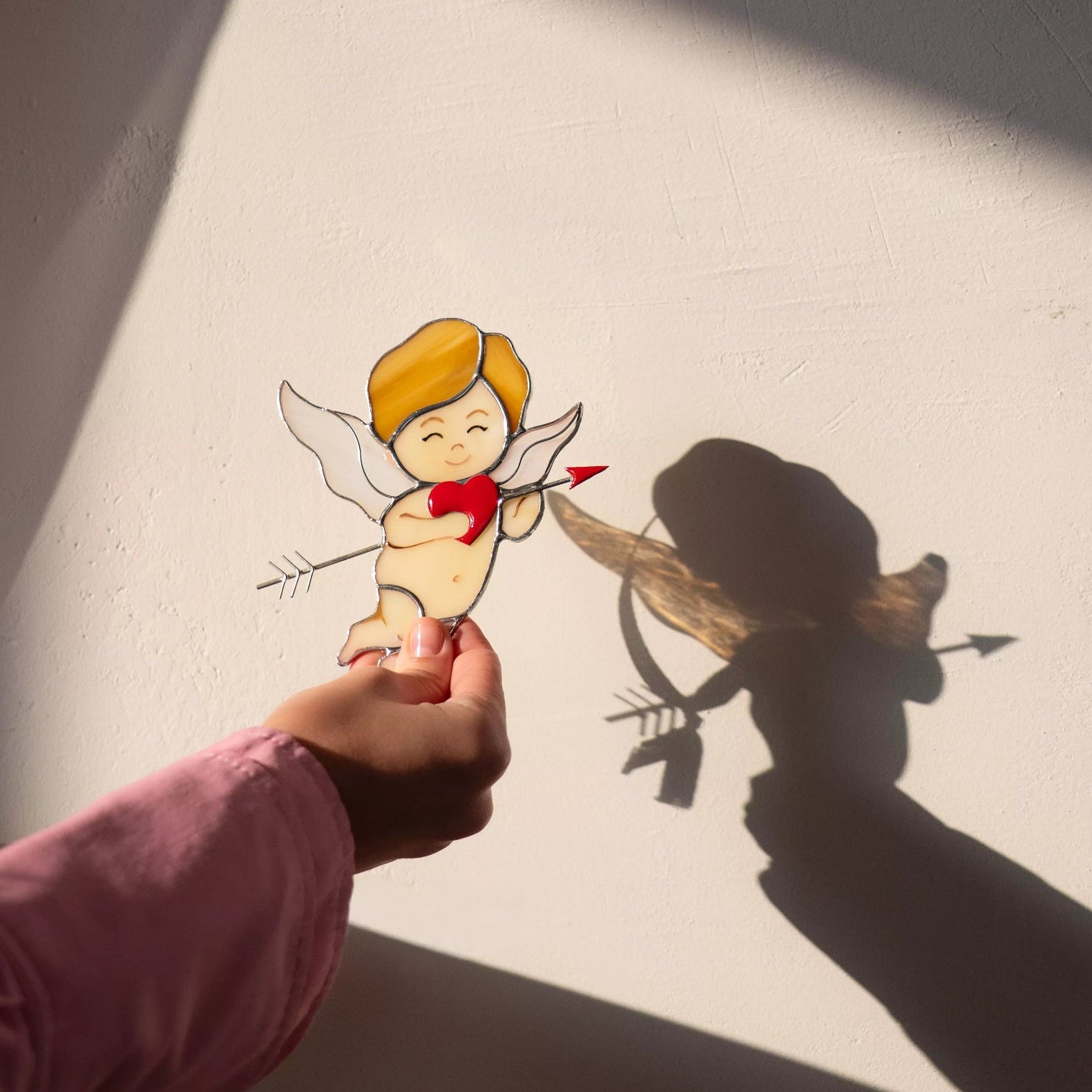 Hand holding a stained glass cupid suncatcher in sunlight, casting a cupid-shaped shadow on the wall.