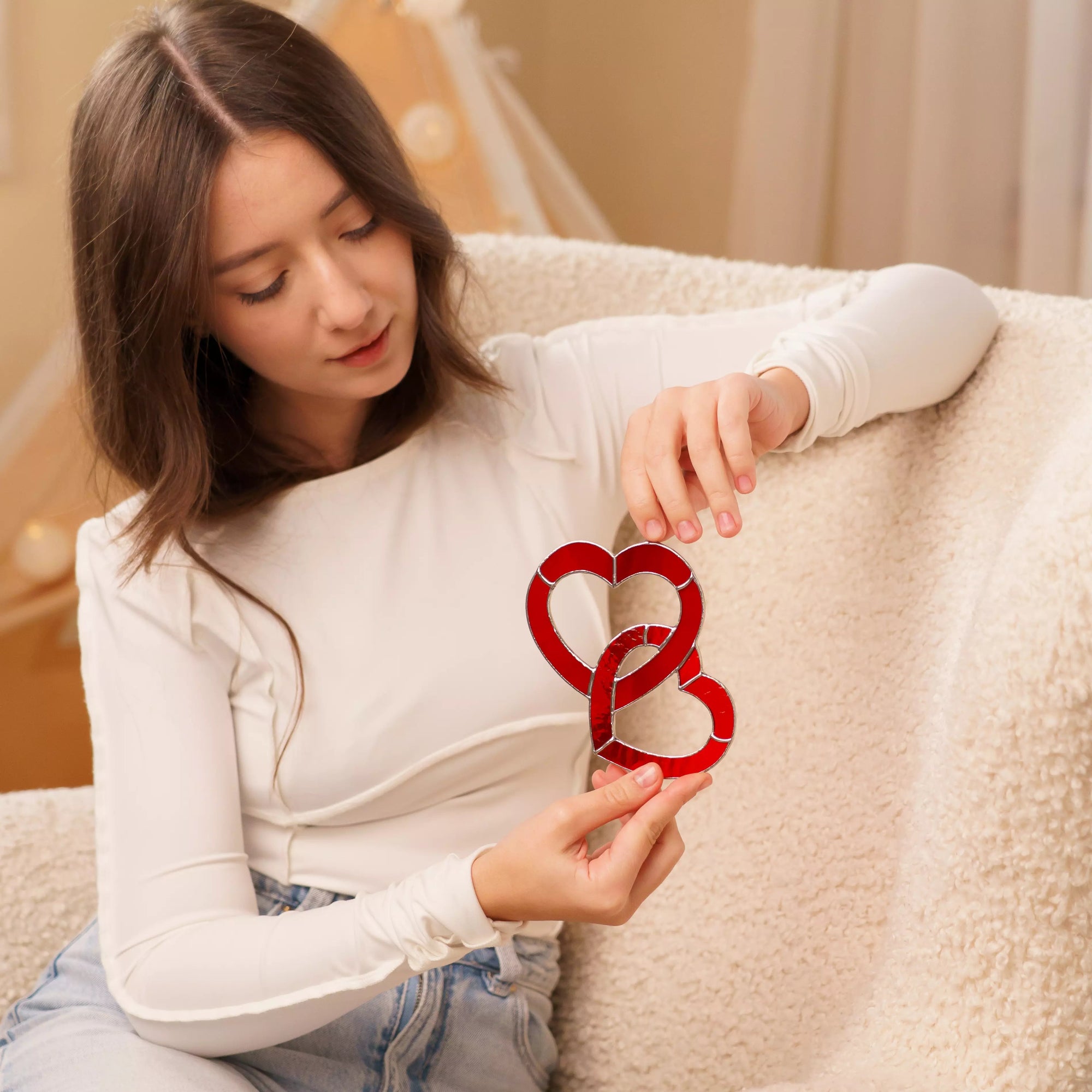 Woman holding a red stained glass heart suncatcher in a cozy interior setting.