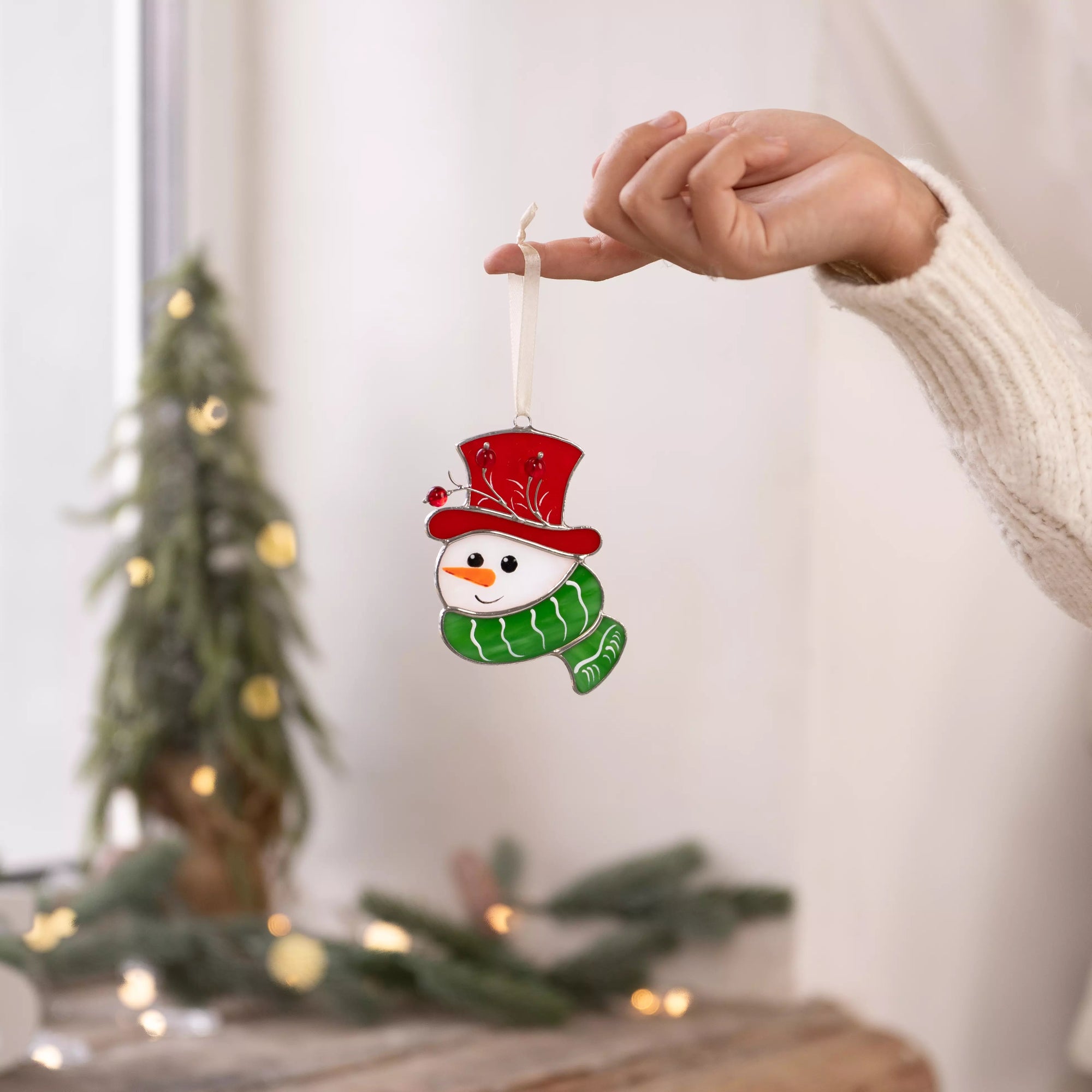 Hand holding a stained glass snowman ornament near a bright window with Christmas tree in the background.