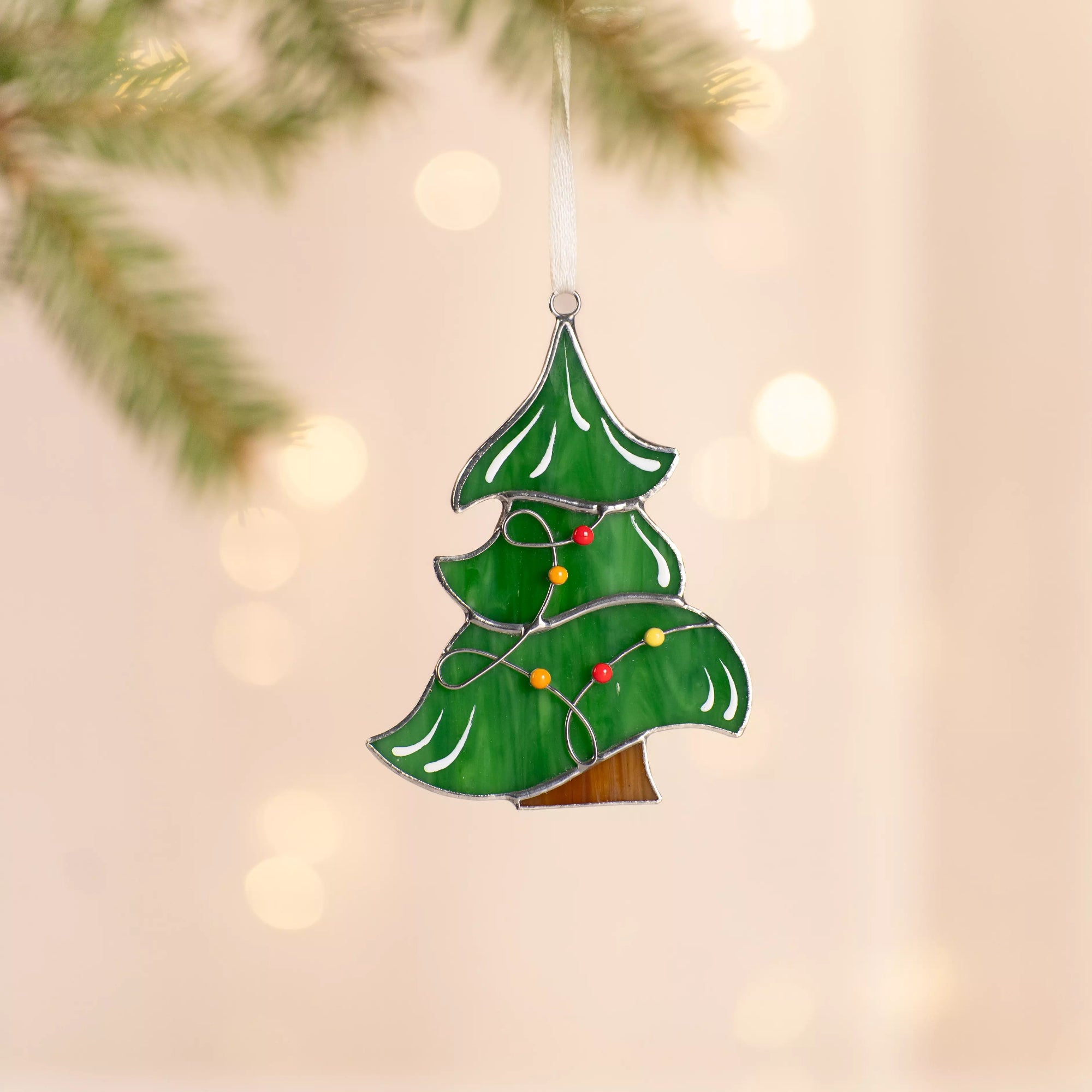 Green stained glass Christmas tree with red and yellow lights on garland hanging near pine branch against a glowing background.
