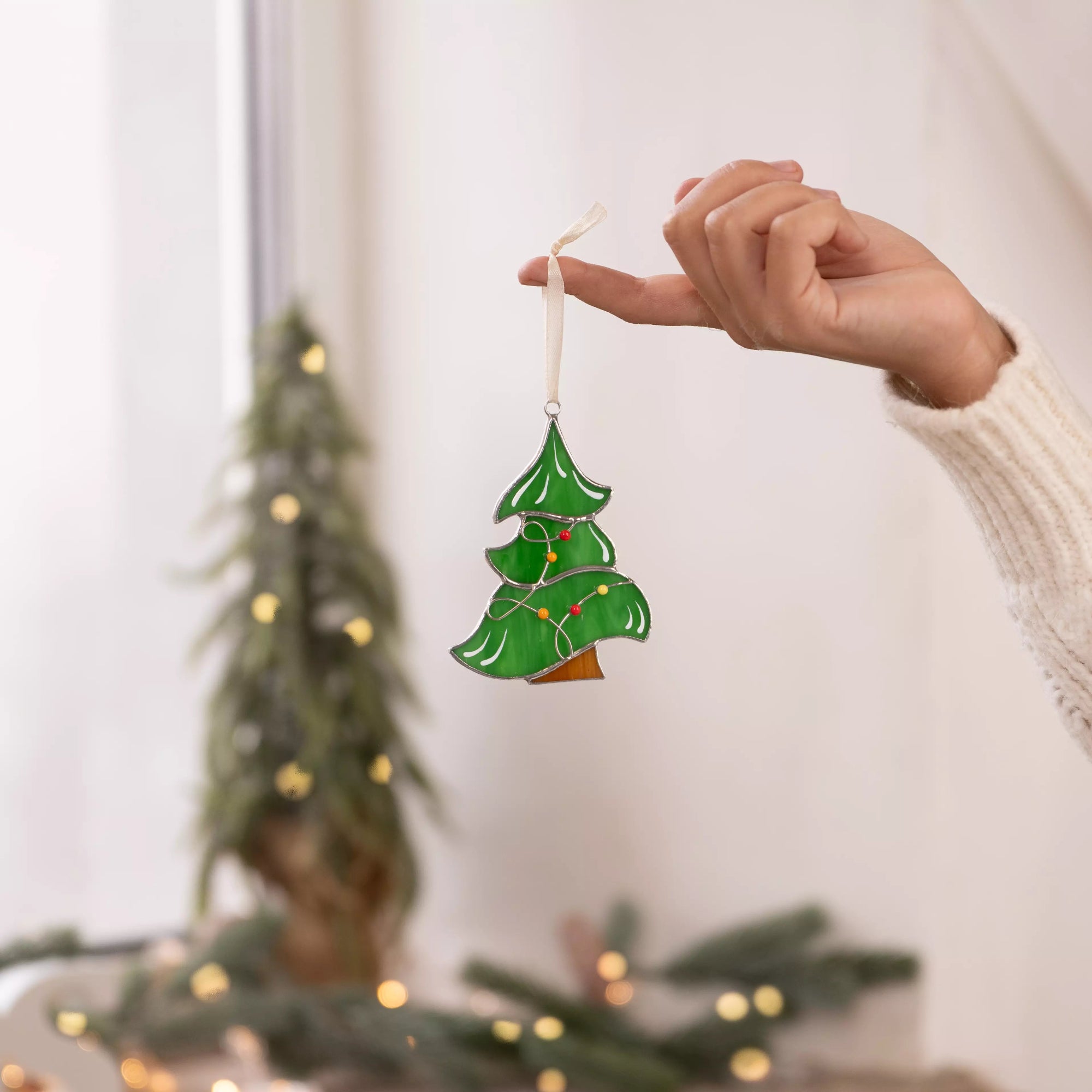 Hand holding a green stained glass Christmas tree ornament near a bright window with soft holiday lights.