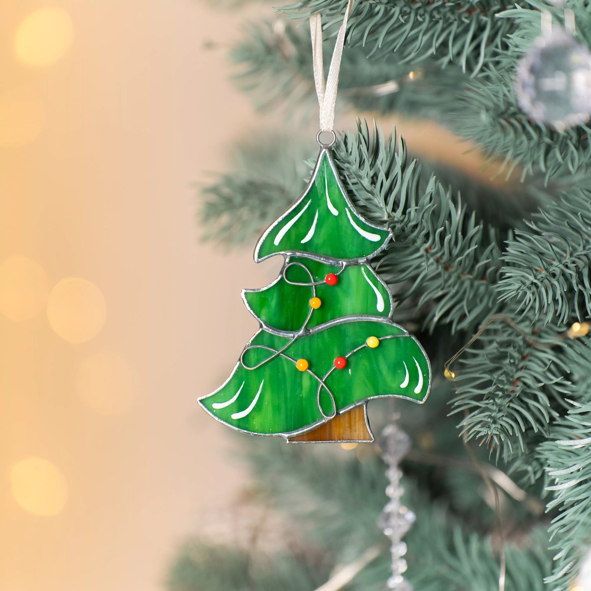 Green stained glass Christmas tree ornament with red and yellow garland beads hanging on a pine branch with warm lights.