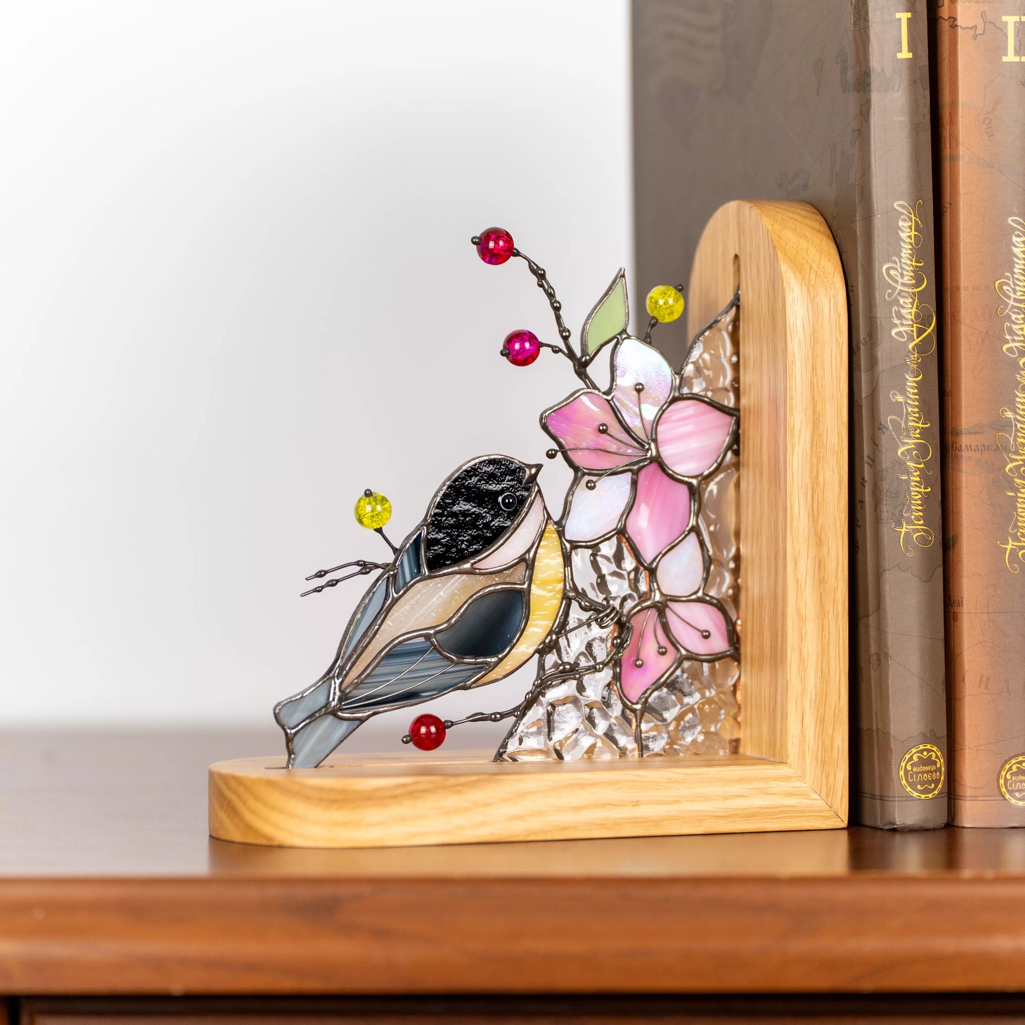Stained glass bird bookend with a chickadee perched among pink flowers, clear textured glass, decorative beads, and a wooden stand on a shelf.
