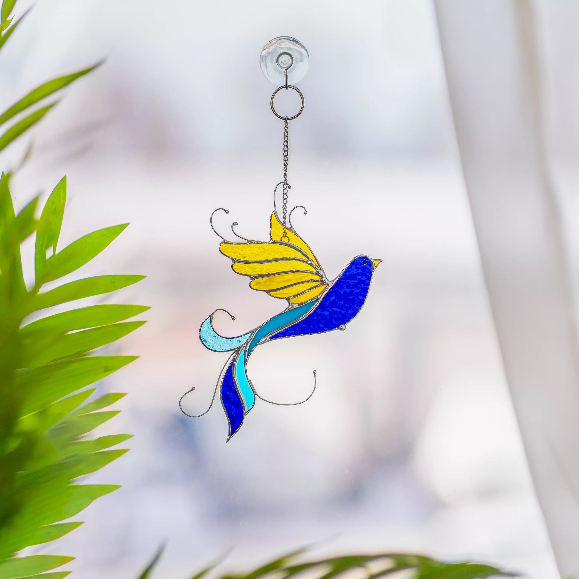 Stained glass bird suncatcher with yellow wings and blue body hanging against a blurred natural background.