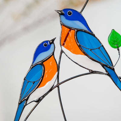 Close-up of two stained glass bluebirds in blue and orange glass perched on a branch with green leaf