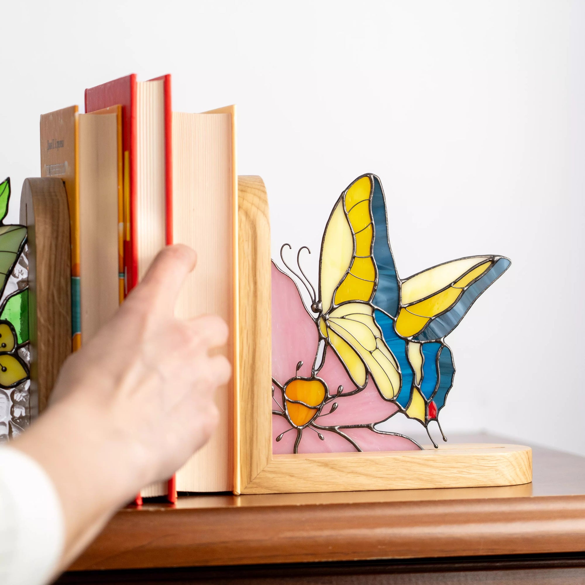 Stained glass butterfly bookend supporting books on a wooden shelf, shown in use while a hand pulls out a book, demonstrating both decorative and functional design.