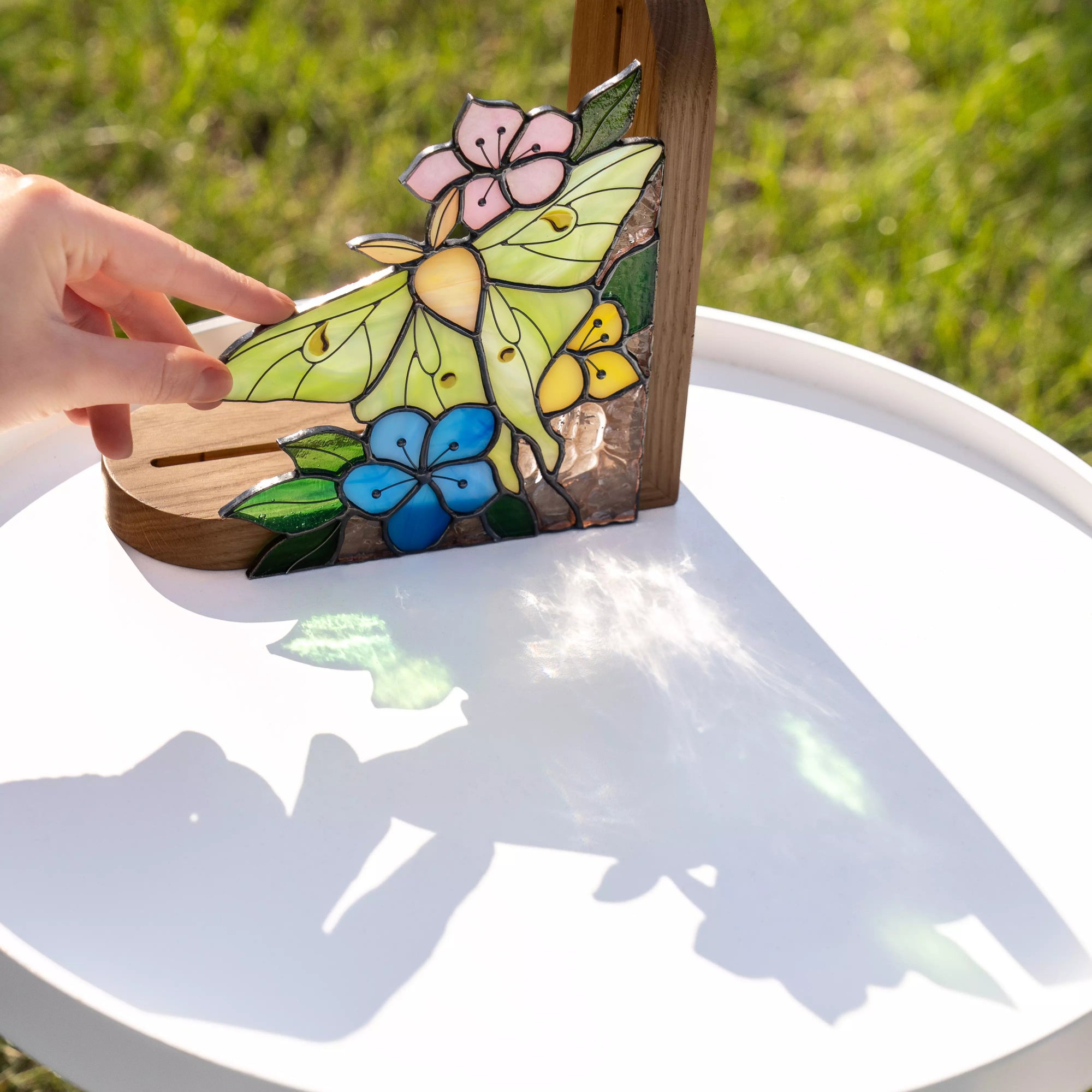 Hand touching stained glass luna moth bookend outdoors, sunlight creating soft reflections and shadows