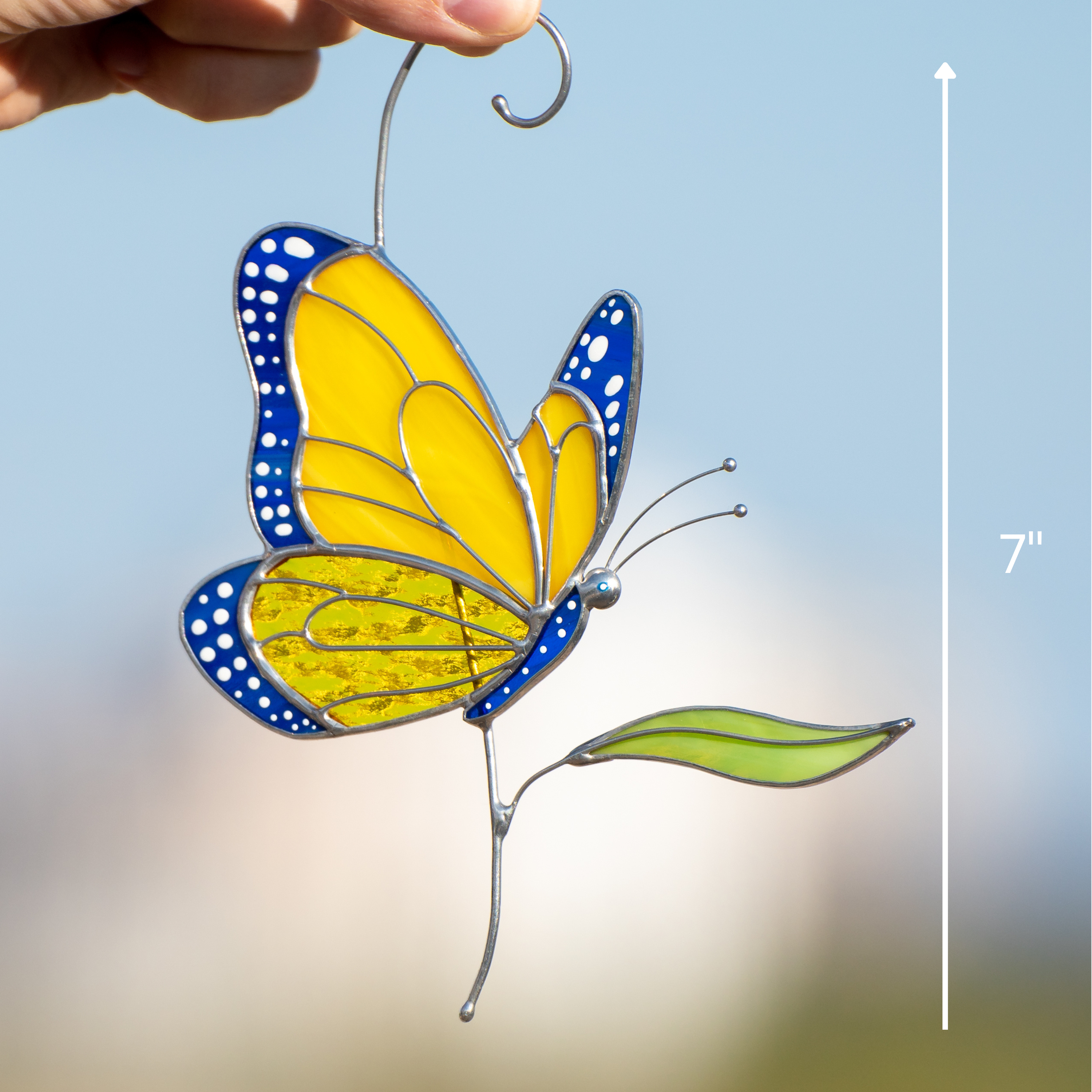 Stained glass butterfly held by hand, yellow wings with blue details, green leaf, and size marking 7 inches.