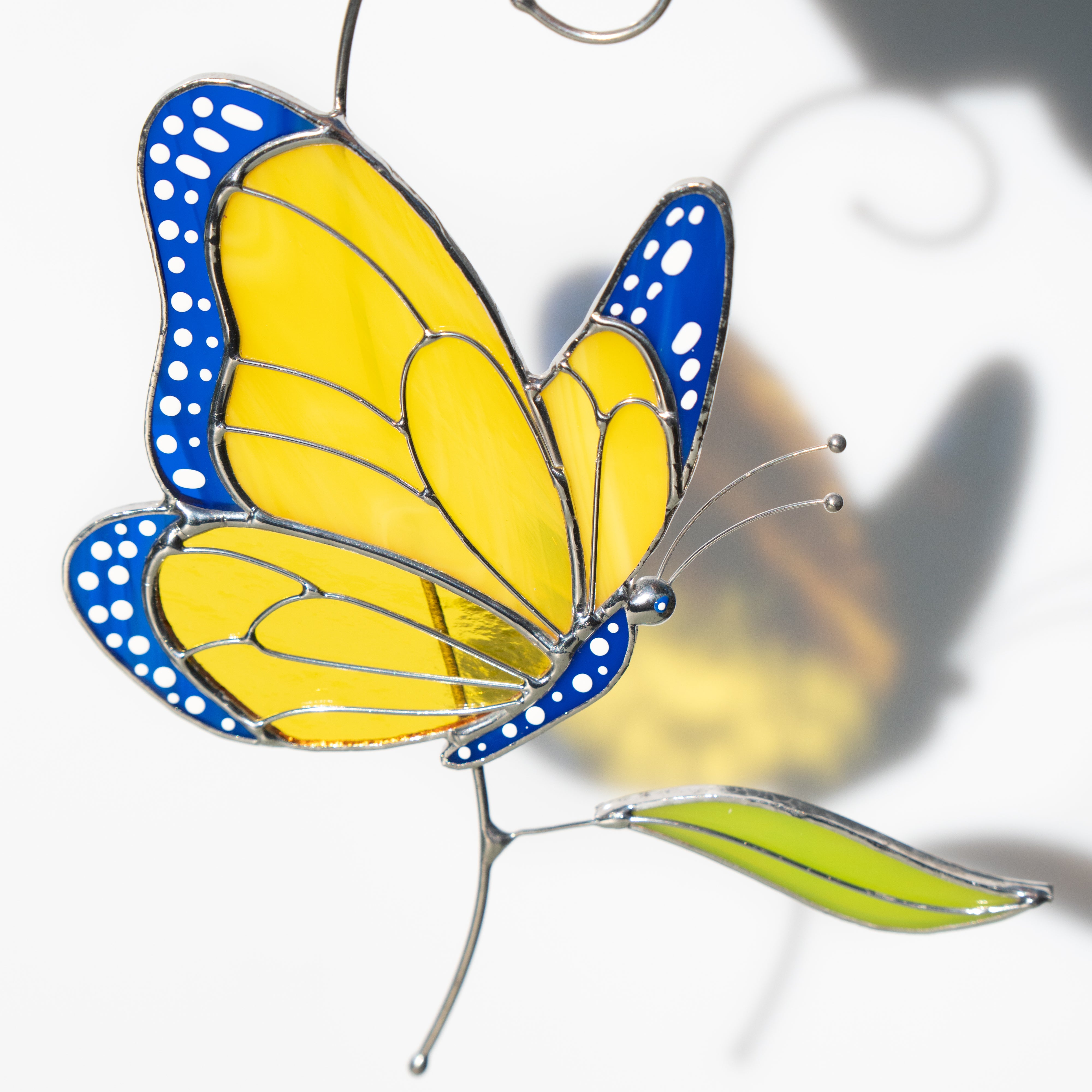 Yellow and blue stained glass butterfly with a green glass leaf casting a delicate shadow on a white background.