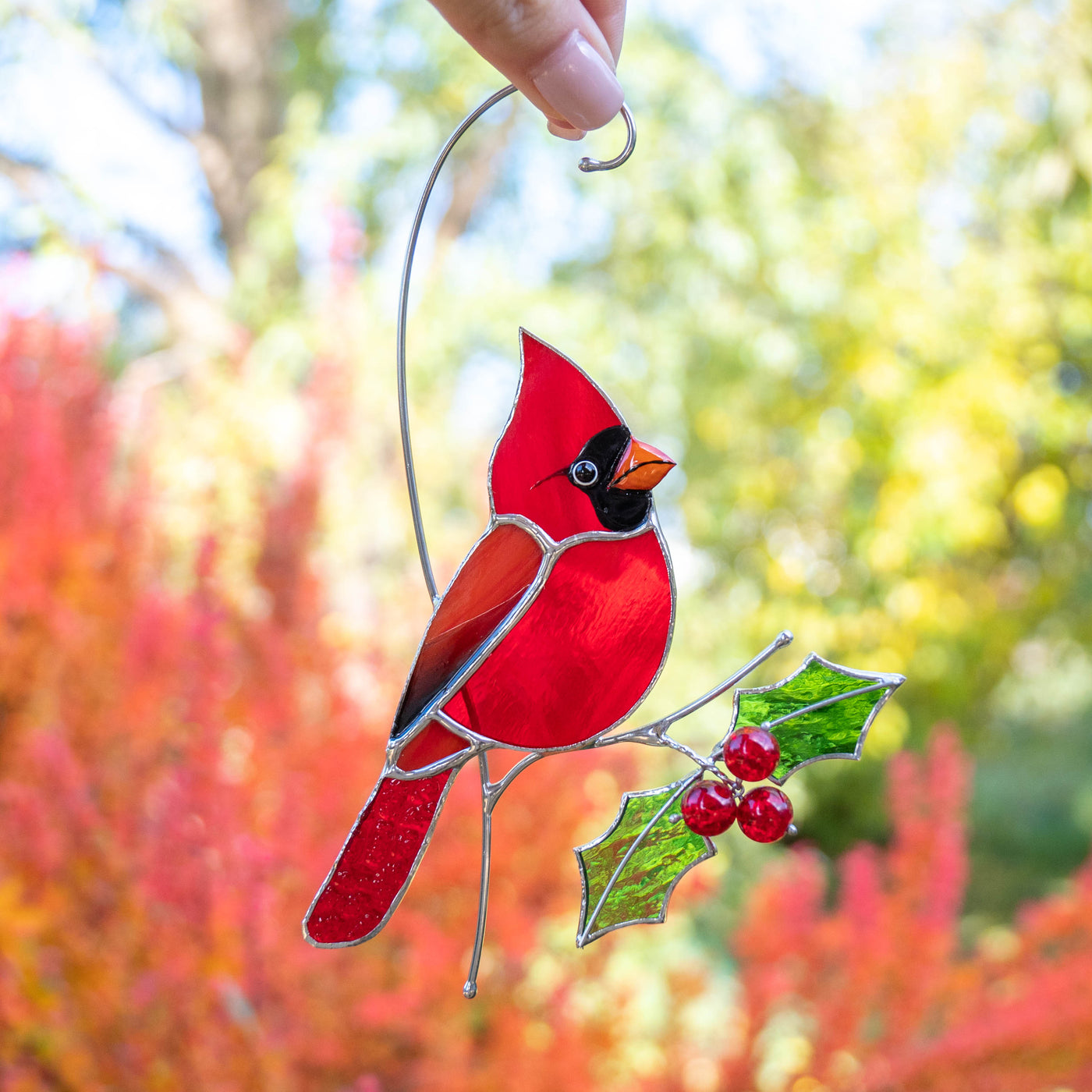 Red stained glass cardinal with green holly leaves held by hand, bright autumn background visible.