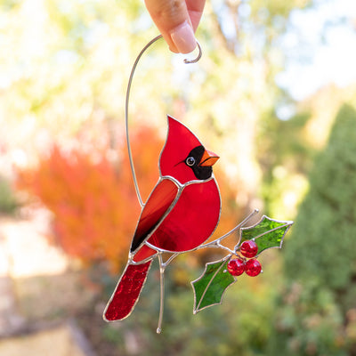 Stained glass cardinal with vibrant red glass and green holly leaves held by hand against fall colors.