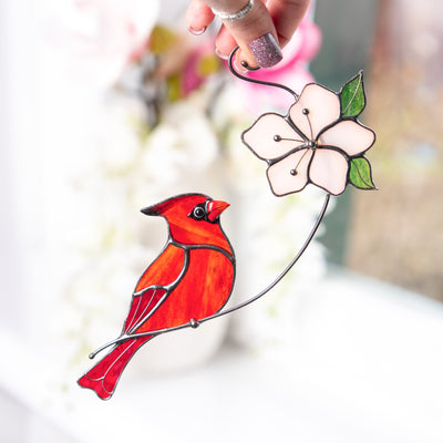 Hand holding a stained glass red cardinal perched next to a delicate pink flower with green leaves.