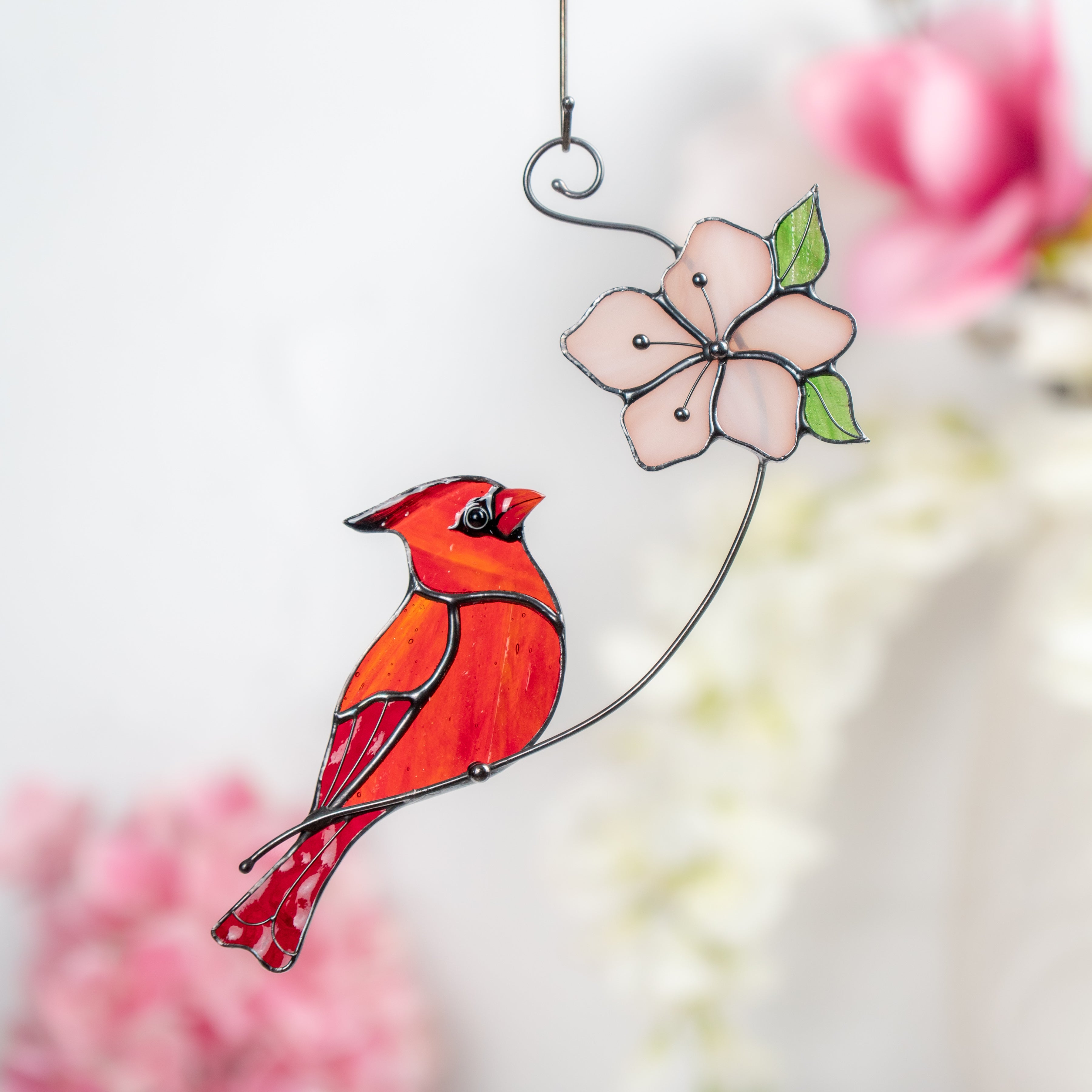 Hand holding a stained glass red cardinal perched next to a delicate pink flower with green leaves.