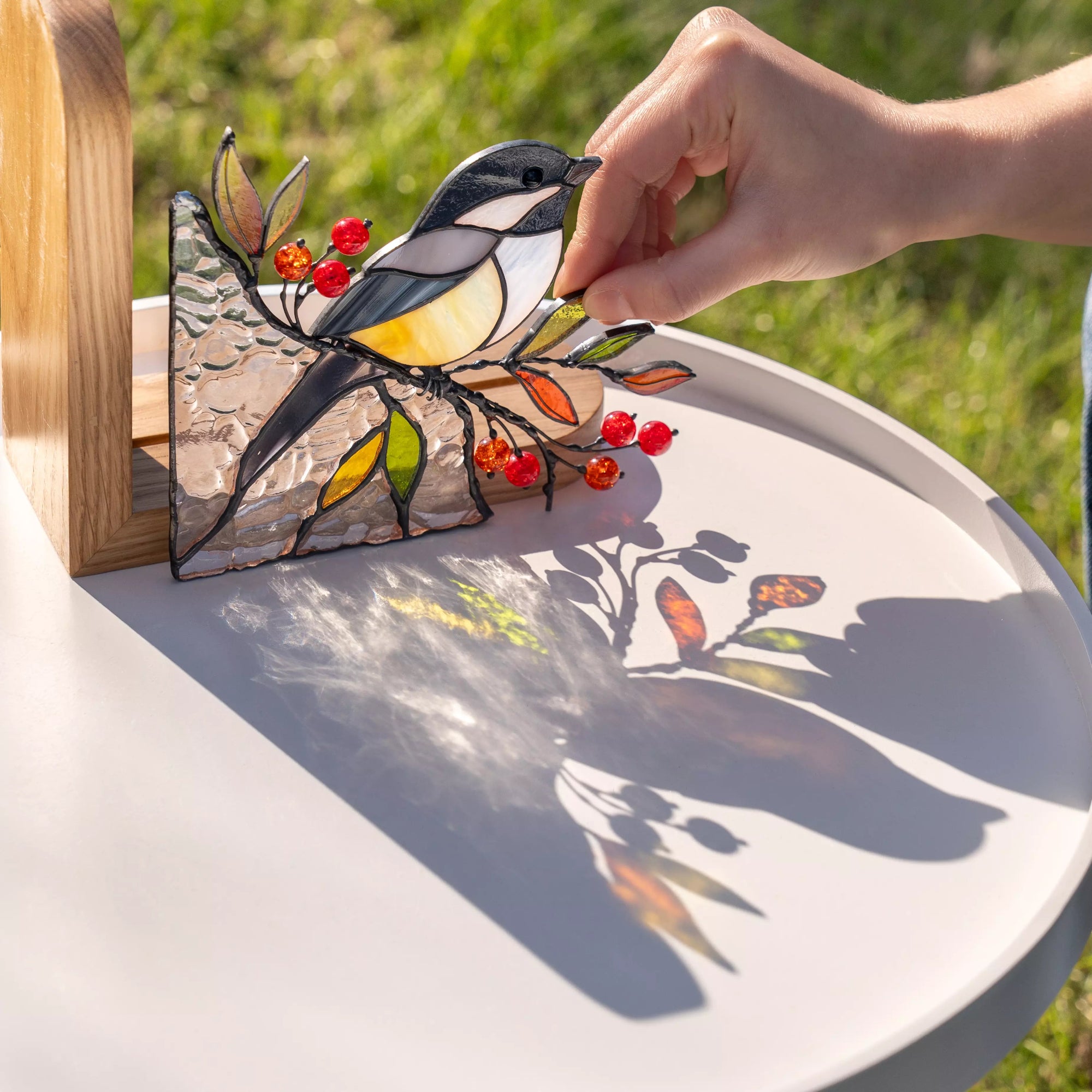Handcrafted stained glass chickadee bird with branch, beads, and leaves, casting colorful reflections on a light surface.