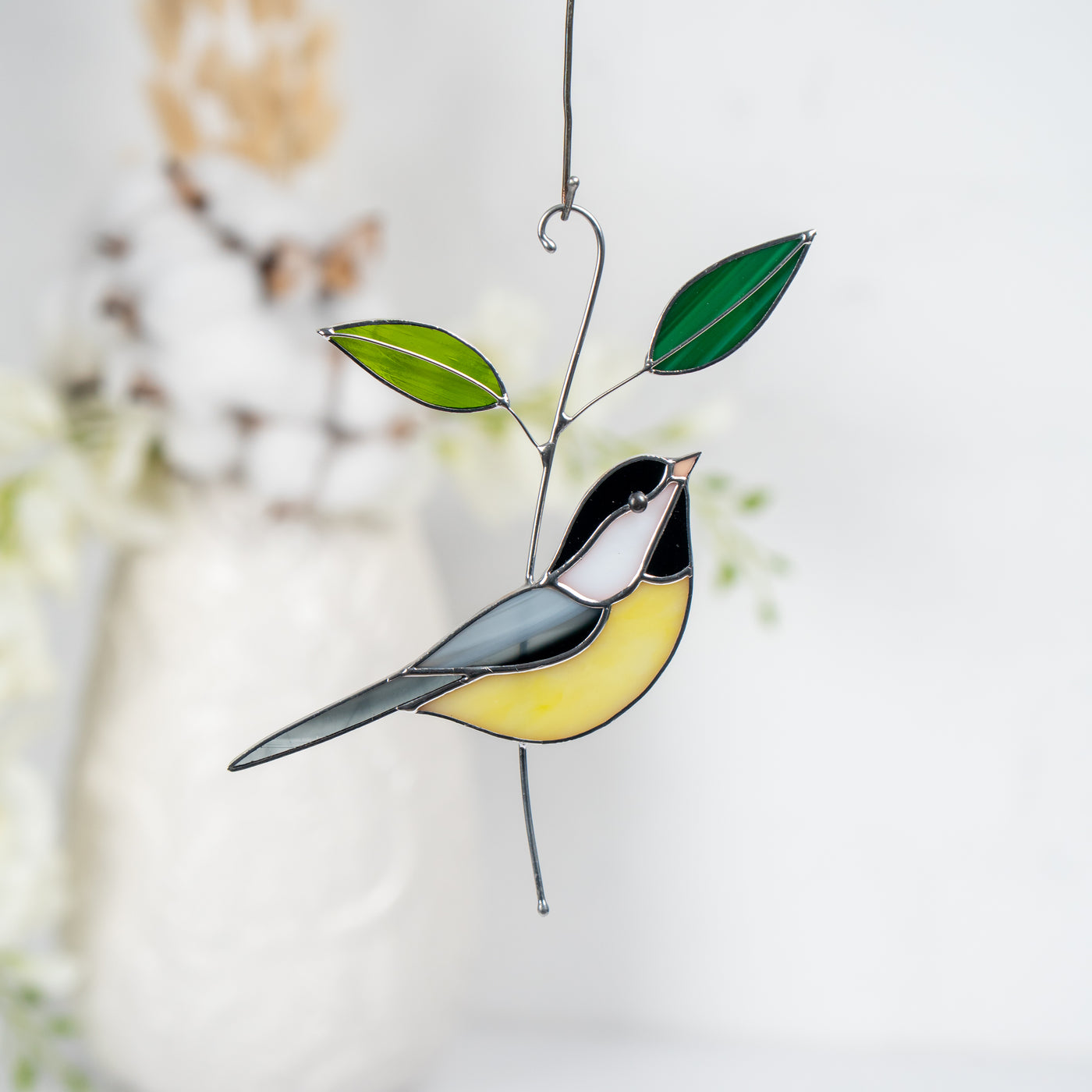 Yellow and black stained glass chickadee with two green leaves, hanging in soft natural light with floral background