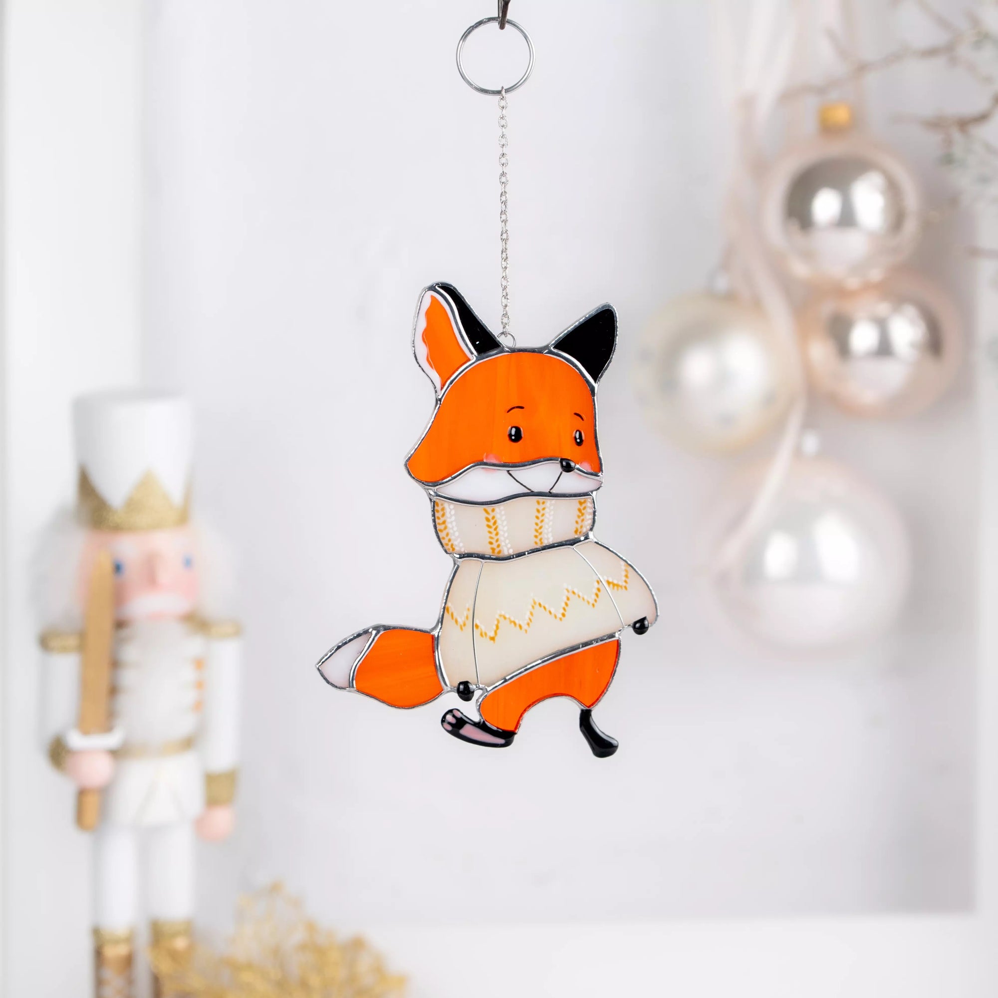 Stained glass suncatcher fox in a sweater hanging against a white background with Christmas decorations.