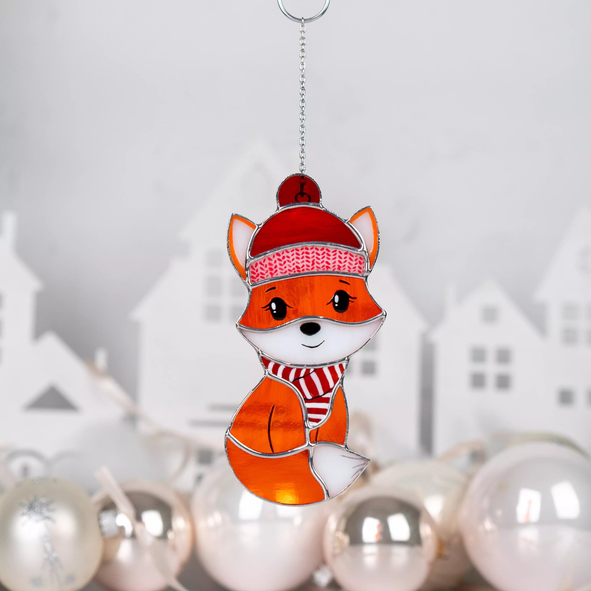 Stained glass ornament featuring a fox wearing a red hat and scarf with Christmas decorations in the background.