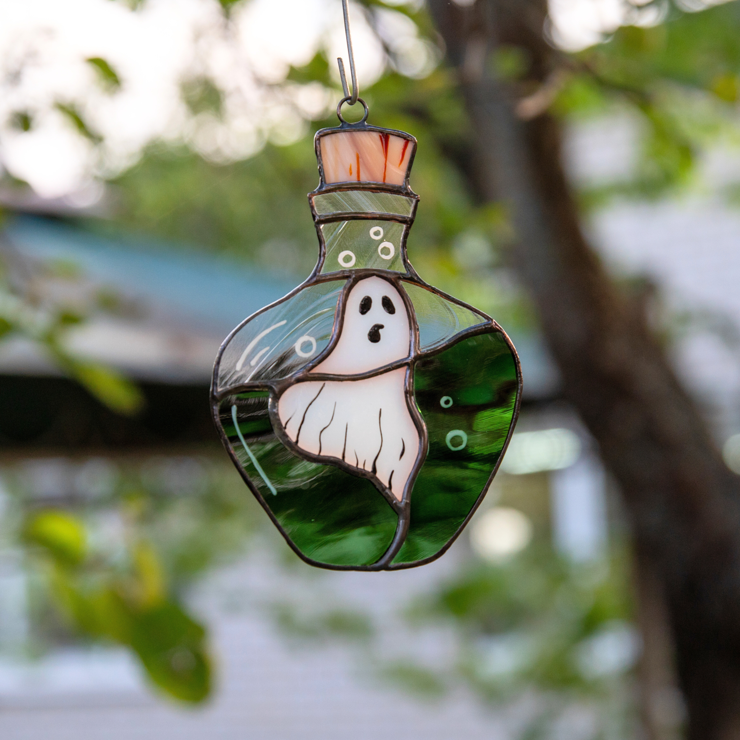 A stained glass ghost suncatcher in a potion bottle shape hanging outside, surrounded by trees and soft evening light.