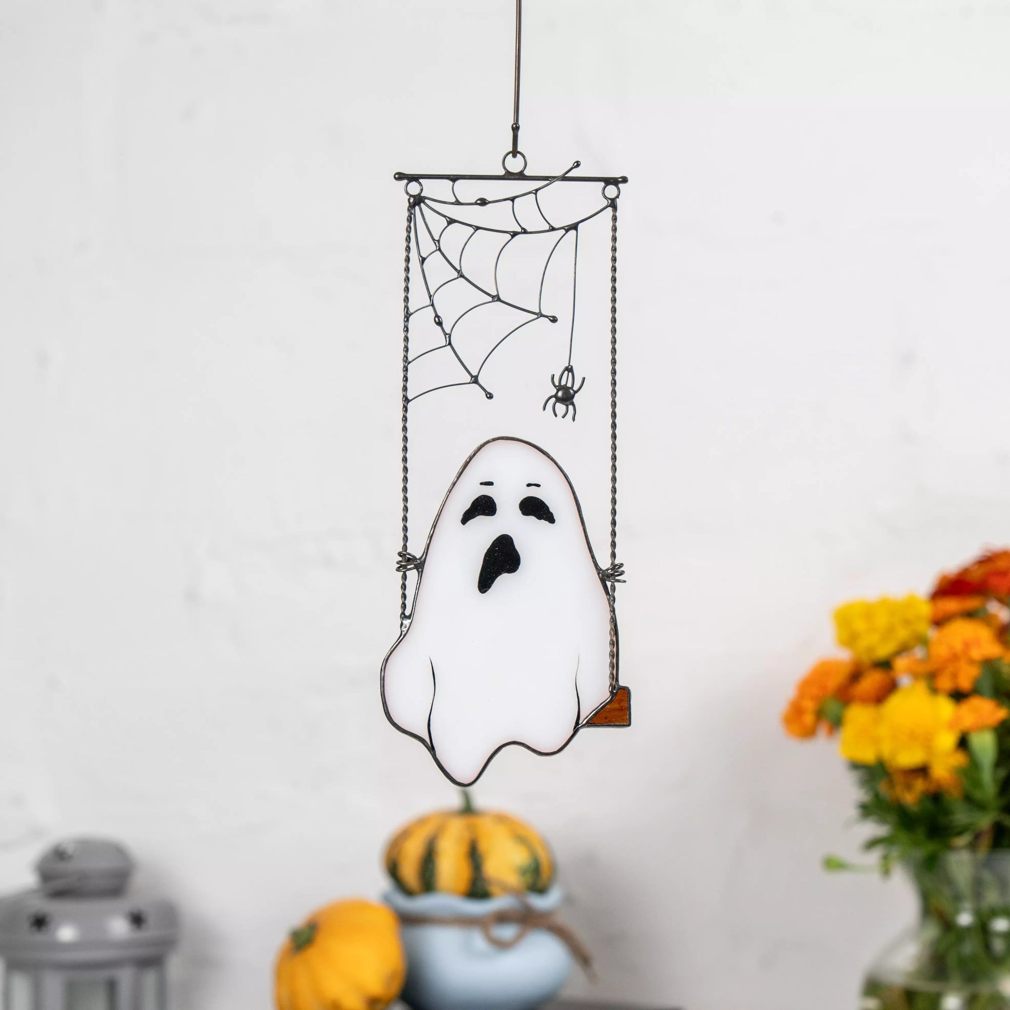 White stained glass ghost on swing under spiderweb hanging indoors with pumpkins