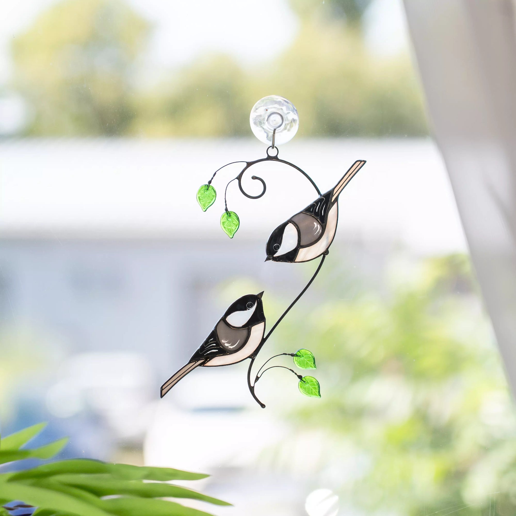 Stained glass window hanging with two birds and green glass leaves against a blurred natural background